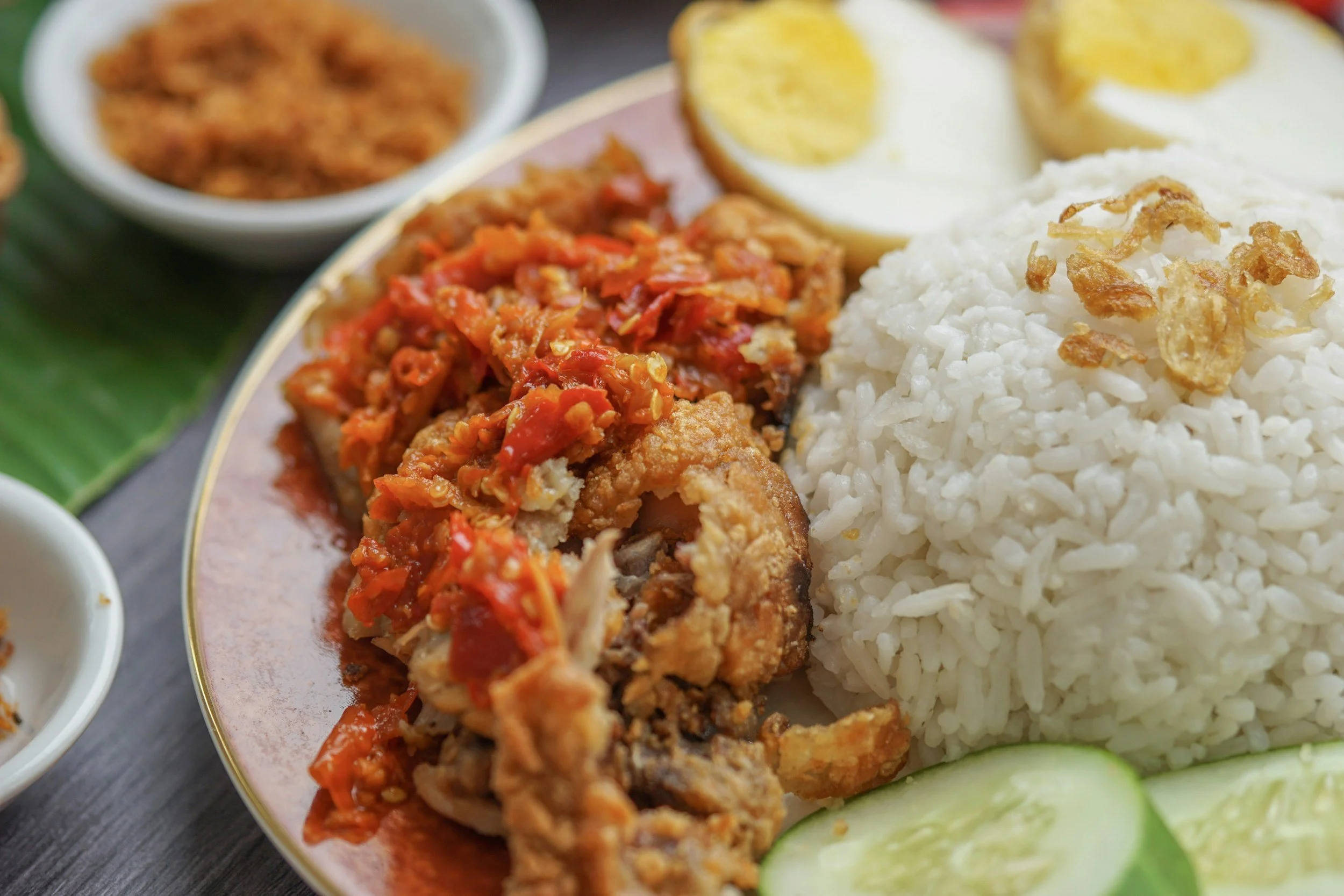 Plate of Indonesian food with fried chicken topped with spicy chili sauce, white rice, half boiled egg, and cucumber slices, with small bowls of additional sauces or sambal in the background.