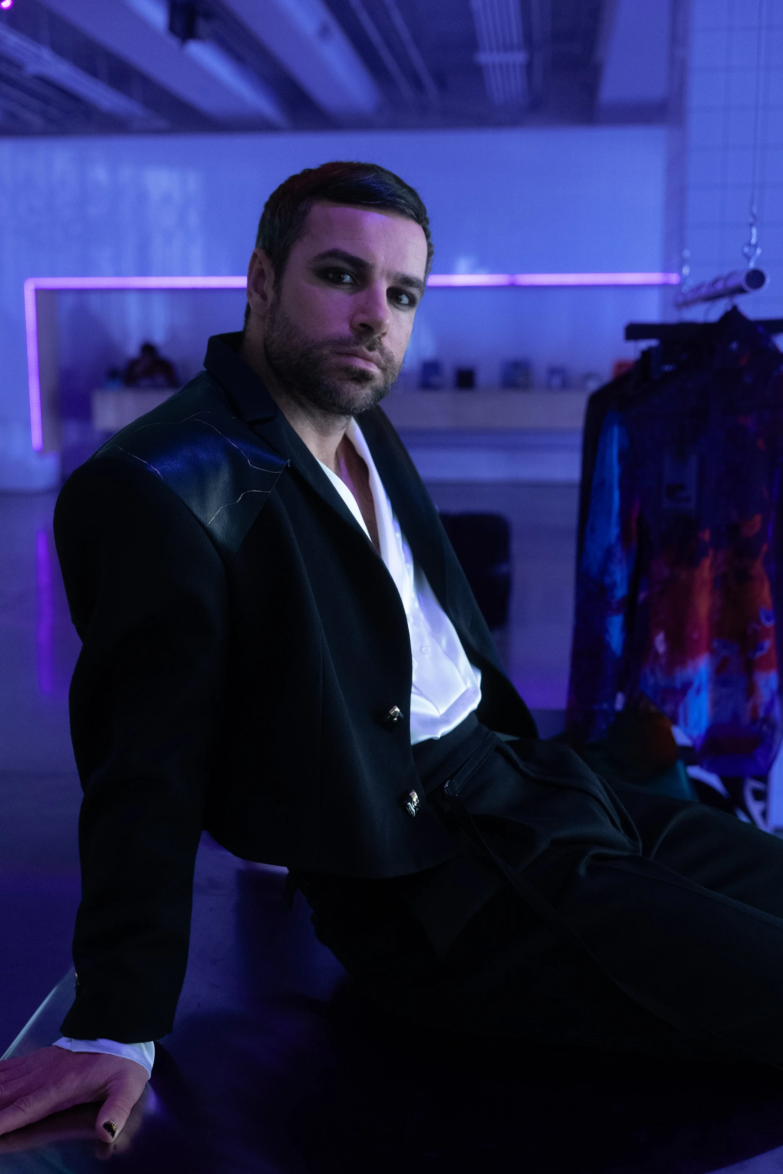 A man with dark hair and a beard sitting on the floor in a neon-lit room, wearing a black blazer and white shirt, with a colorful shirt hanging on a rack beside him.