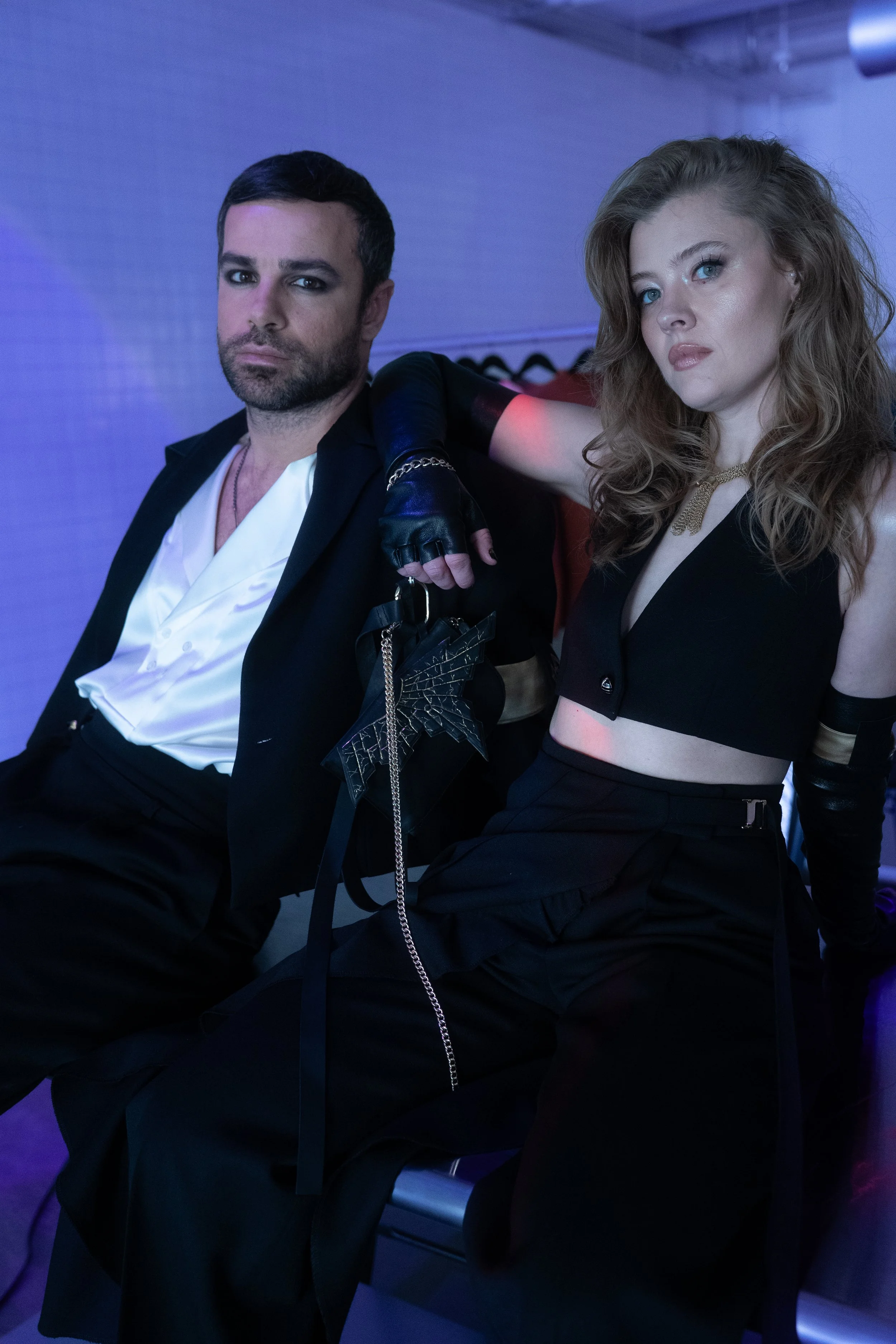 Man and woman dressed in stylish black and white clothing sitting in a modern, dimly lit room with blue and purple lighting.