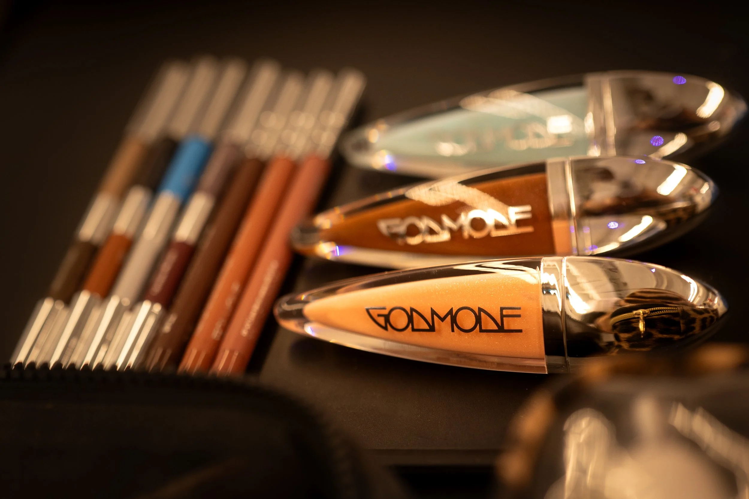 Close-up of three gold-colored makeup compacts with the brand name 'GOLDMONE' on the front, and a blurred row of colored pencils in the background.