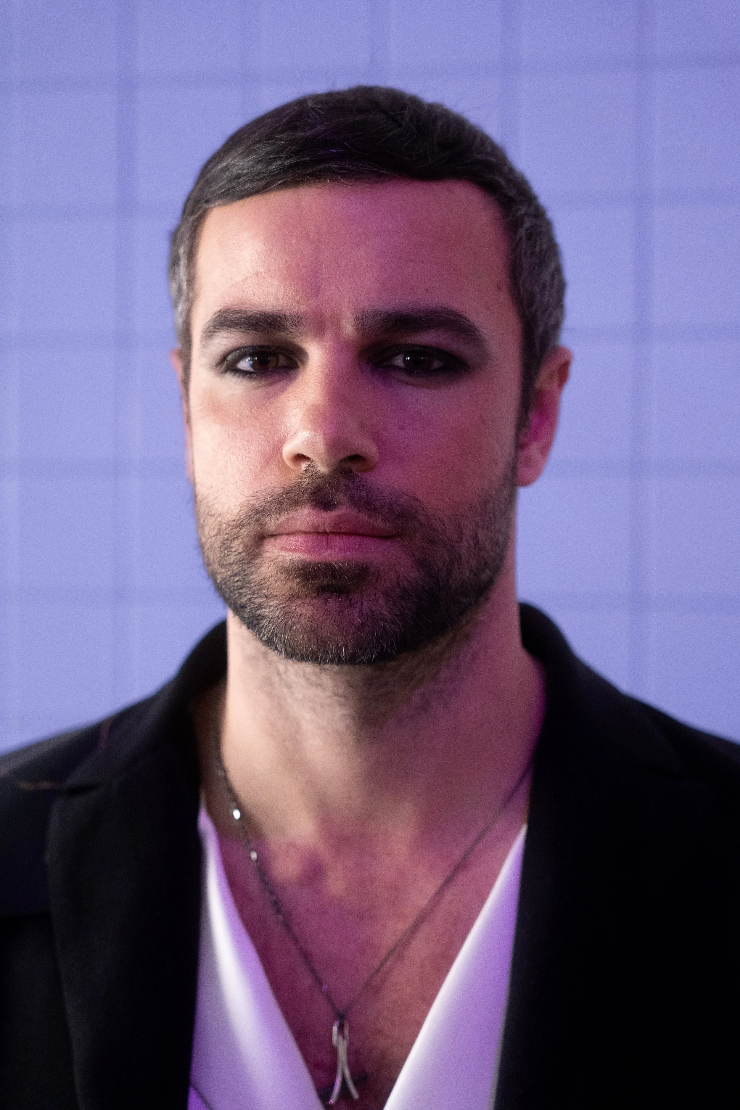 Close-up of a man with dark hair and a beard, wearing a black jacket and a white shirt with a necklace, standing against a tiled wall with purple lighting.