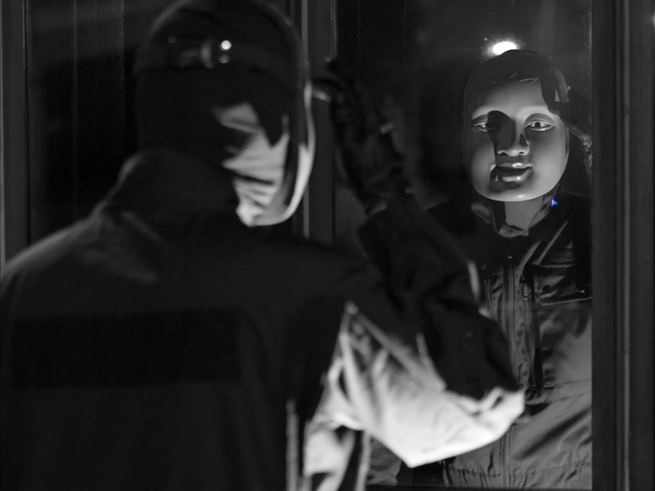 A person wearing a face mask and a cap, looking at a mannequin head with curly hair and painted features behind glass.