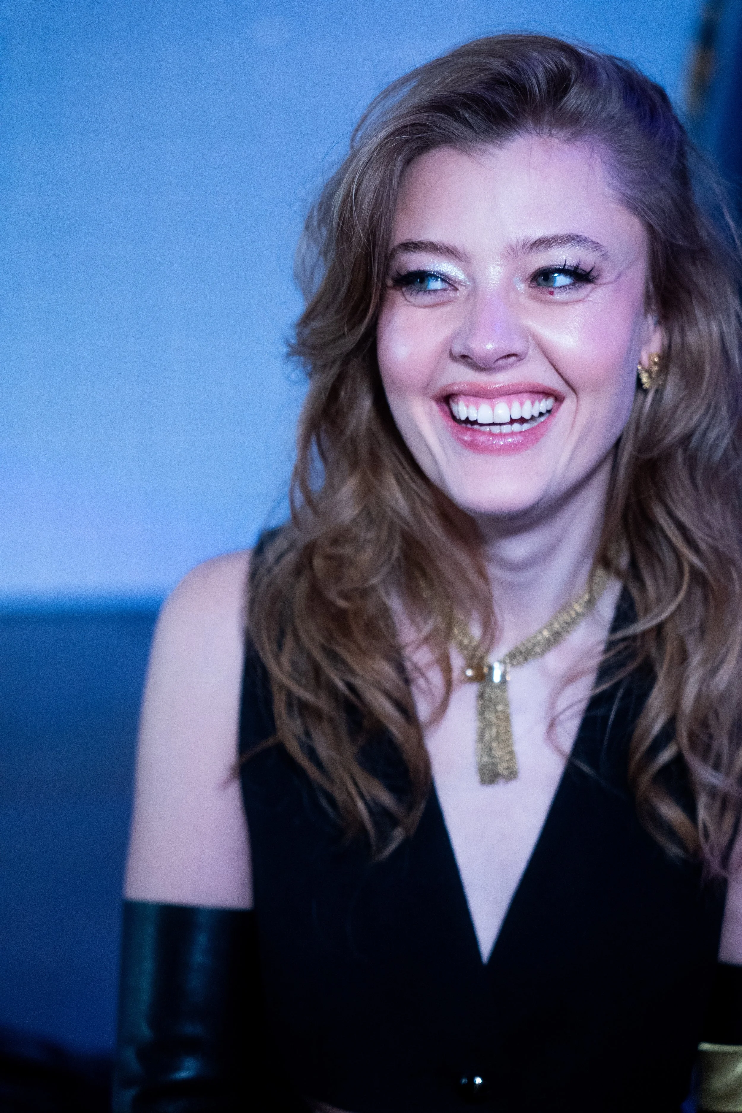 A smiling woman with wavy light brown hair, wearing a black sleeveless top, gold jewelry, and a black arm sleeve, against a blue background.