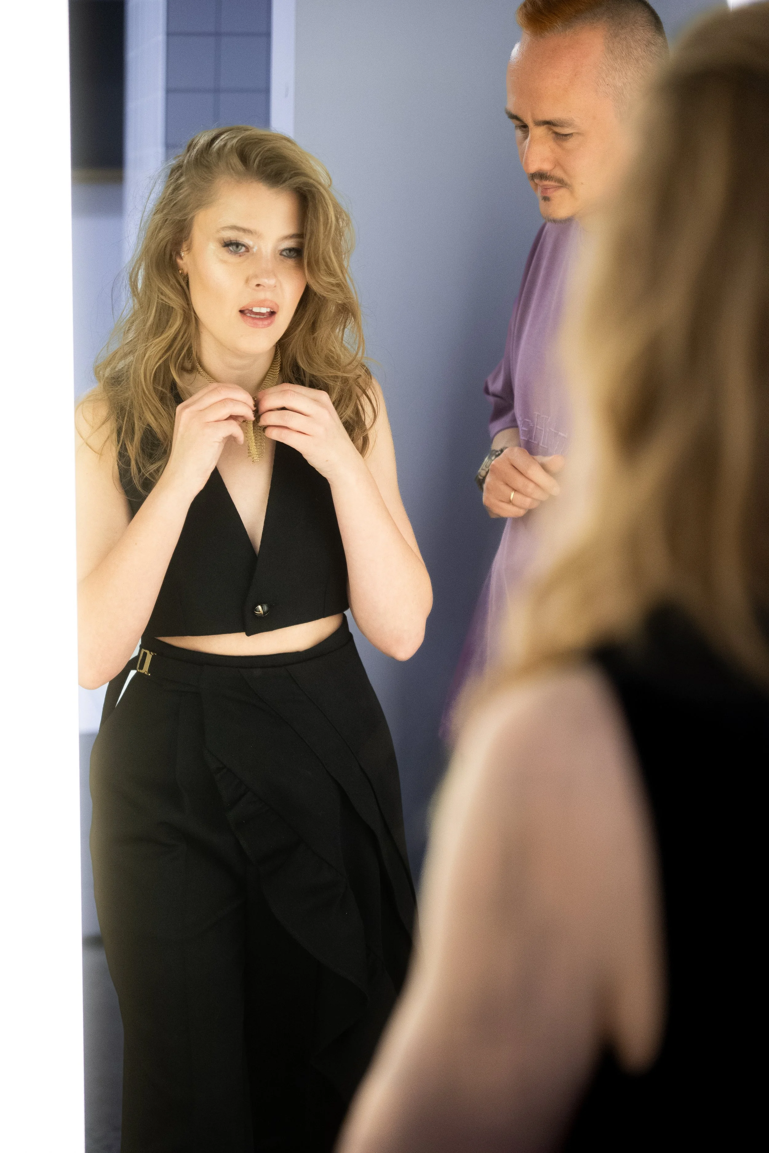 A woman with wavy blonde hair wearing a black sleeveless crop top and high-waisted black pants, talking and gesturing with her hands while a man in a lavender shirt looks on, in an indoor setting.
