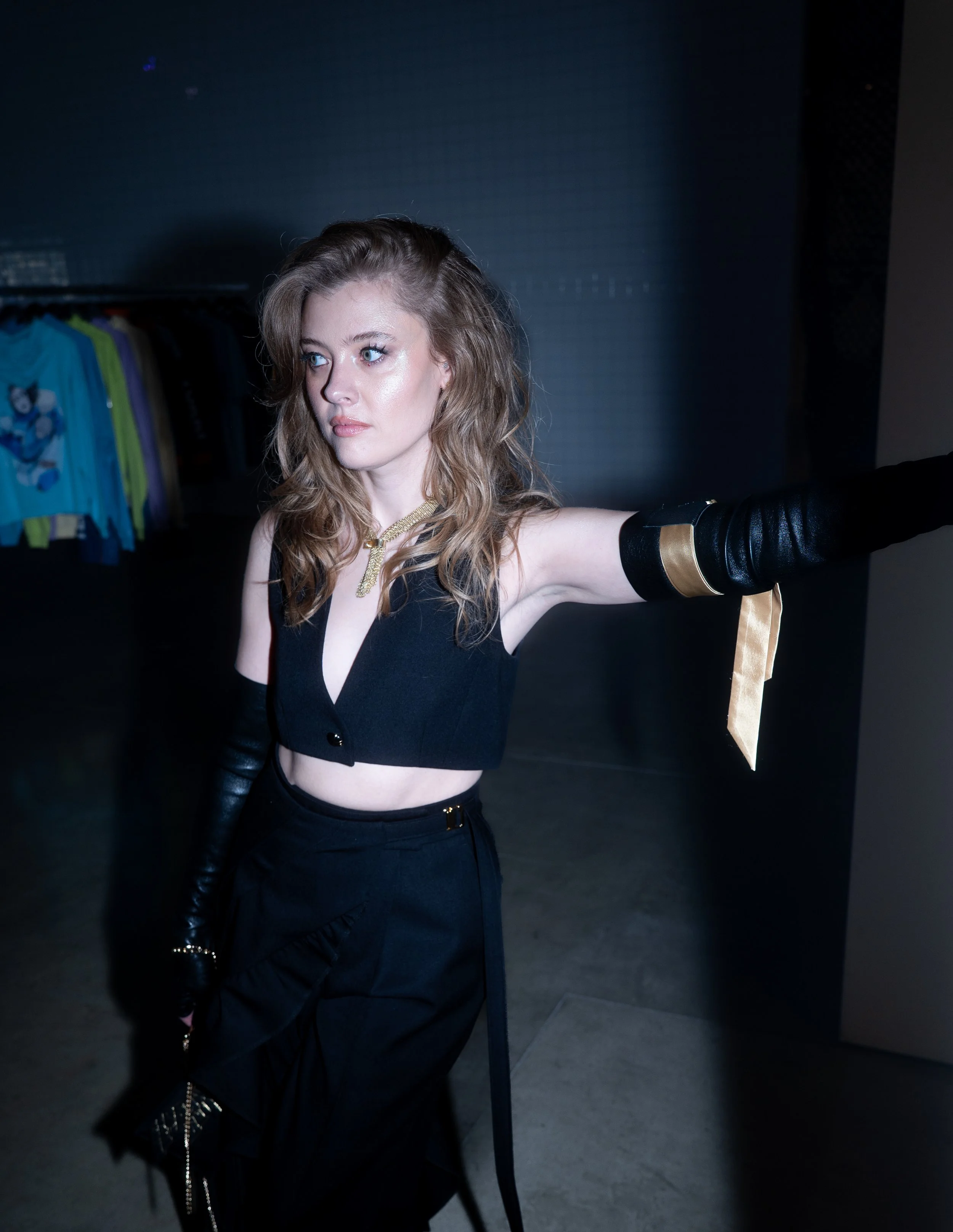 Young woman with light brown, wavy hair and fair skin posing in a dark room with a clothing rack in the background, wearing a black sleeveless top, black pants, and long black gloves with gold accents.