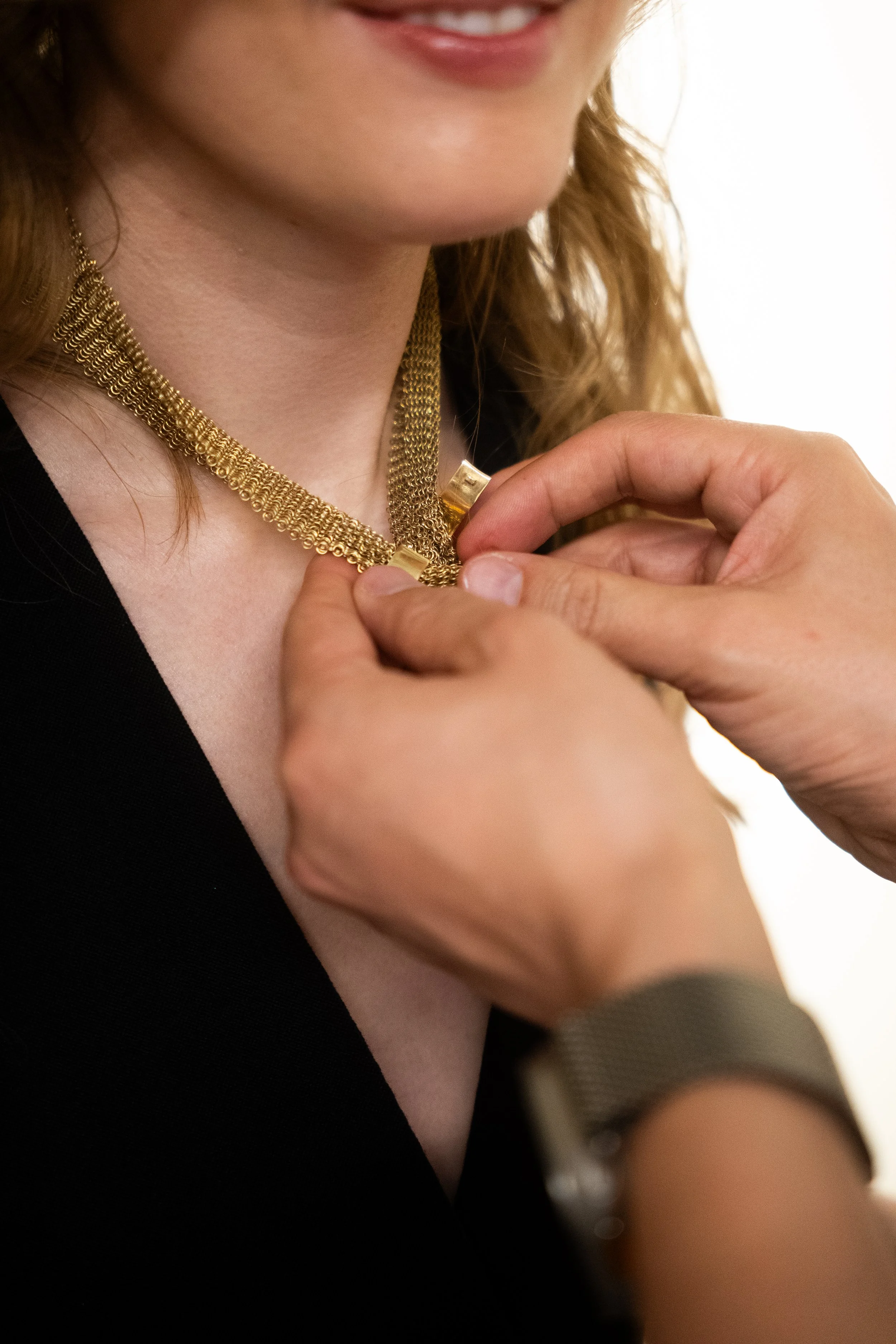A person is putting on a gold necklace with a clasp, while another person holds the necklace in place. The person wearing the necklace has a slight smile, fair skin, and is dressed in black.