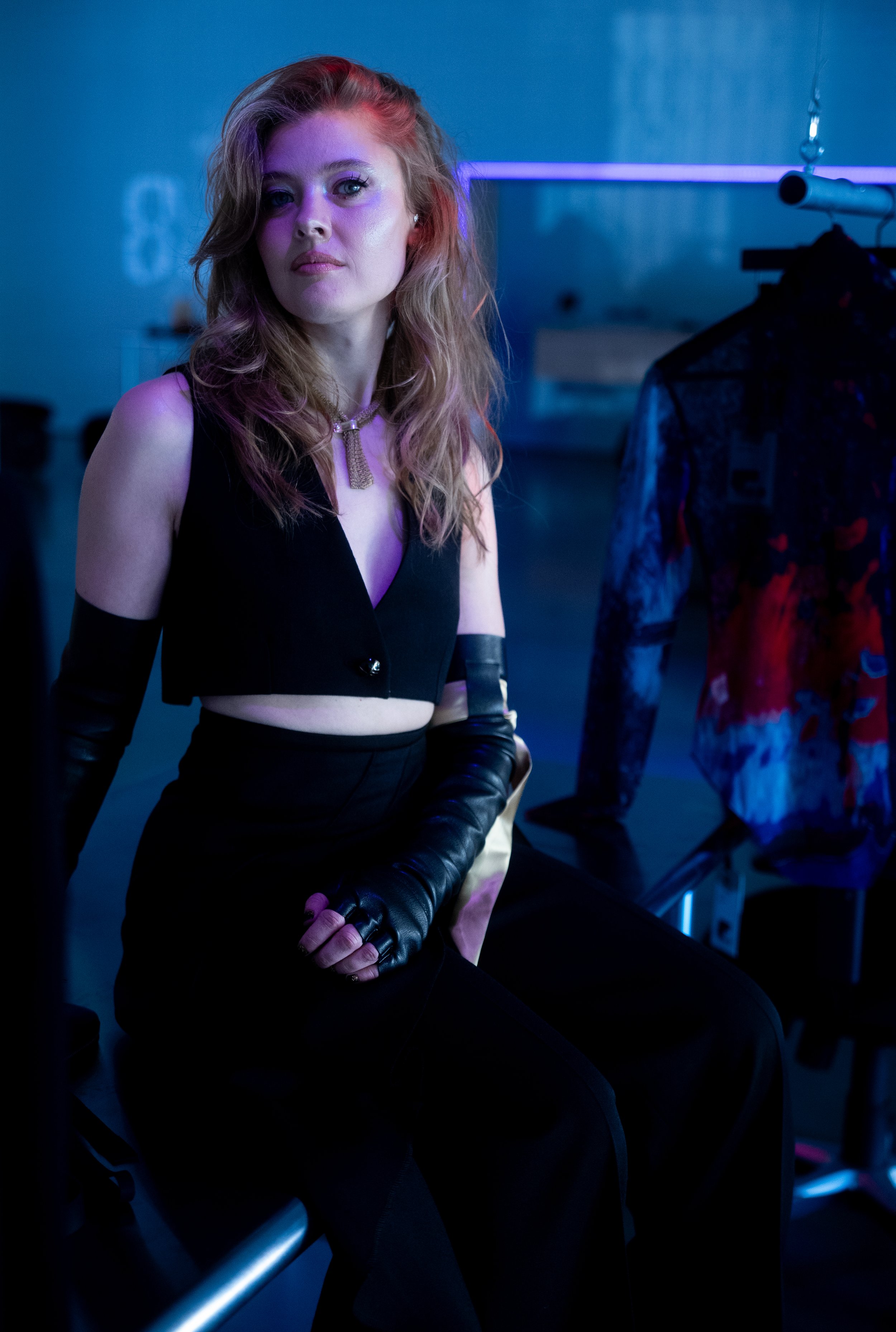 A woman with wavy blonde hair seated in a dimly lit room with blue and purple lighting, wearing a black sleeveless crop top, long black gloves, and a necklace.