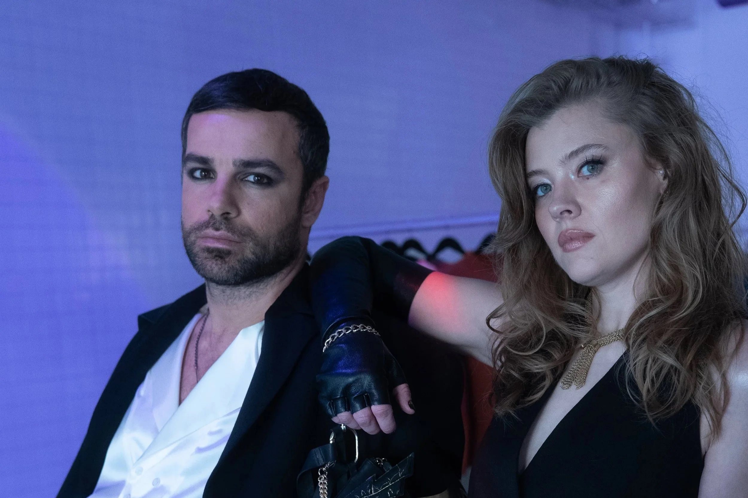 A man and woman with serious expressions standing close together in a dimly lit setting. The man has dark hair, facial hair, and is wearing a suit with a white shirt. The woman has wavy light brown hair, blue eyes, and is wearing a black top with a gold necklace. She has her arm resting on the man's shoulder.