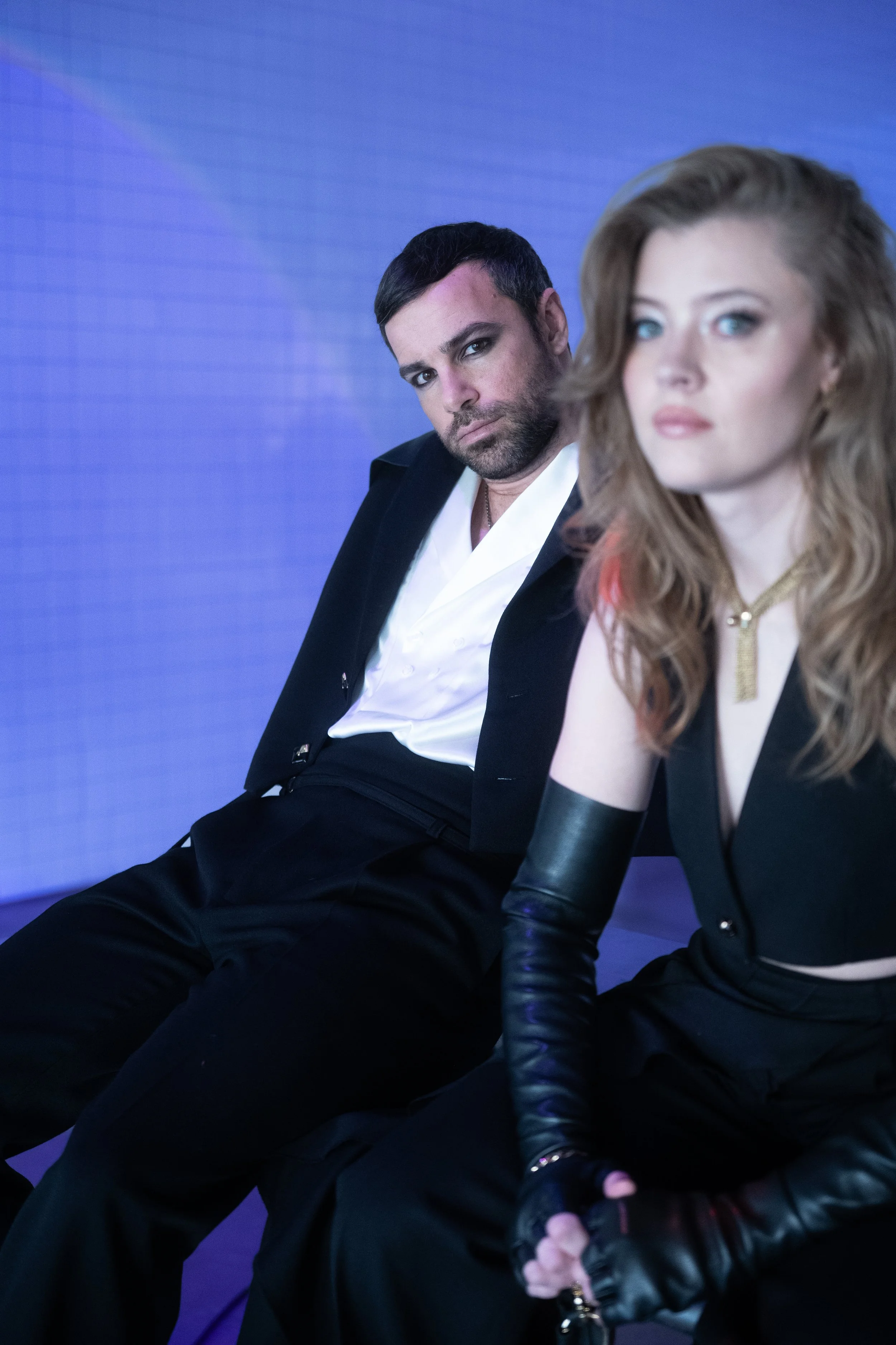 A man and a woman sitting against a blue background, both dressed in dark clothing, with the woman wearing arm-length gloves and a necklace.