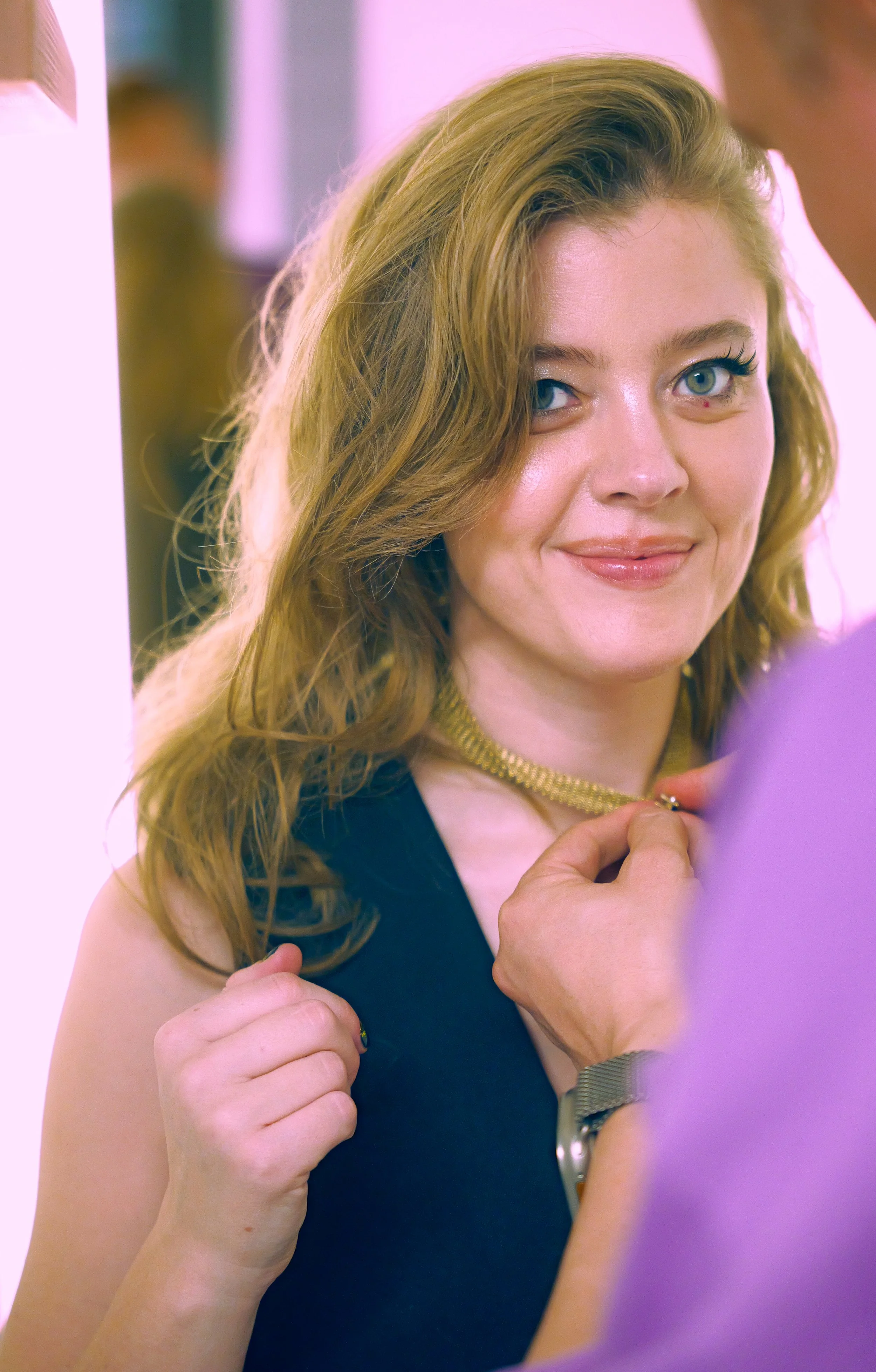 Woman with wavy red hair smiling as a man places a gold necklace around her neck.