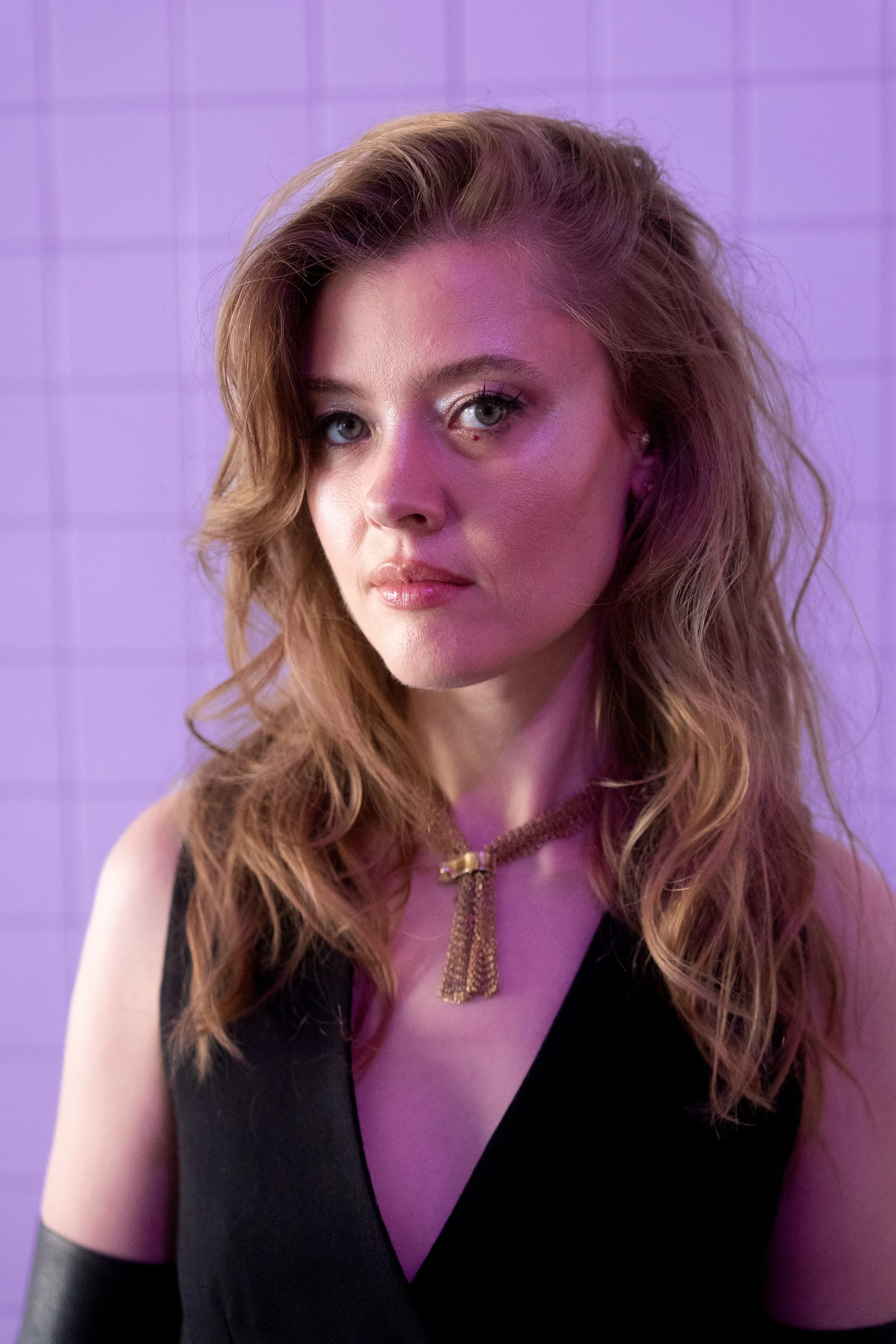Portrait of a woman with wavy, red hair, wearing a black top, and a gold necklace, against a purple tiled background.