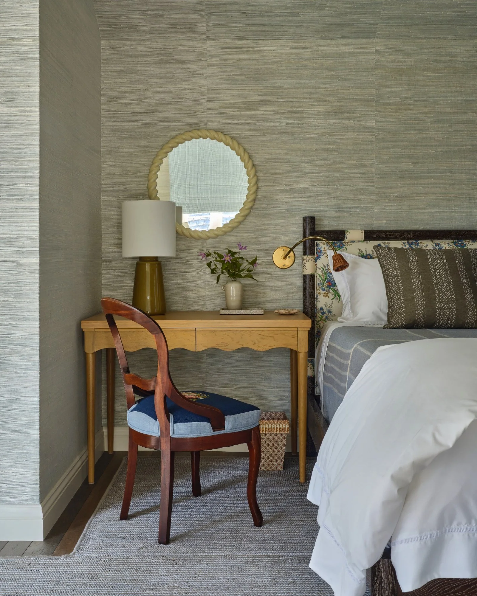 A bedroom corner with a bed, wooden desk, vintage-style chair, mirror, lamp, potted plant, and decorative wall-mounted lamp.