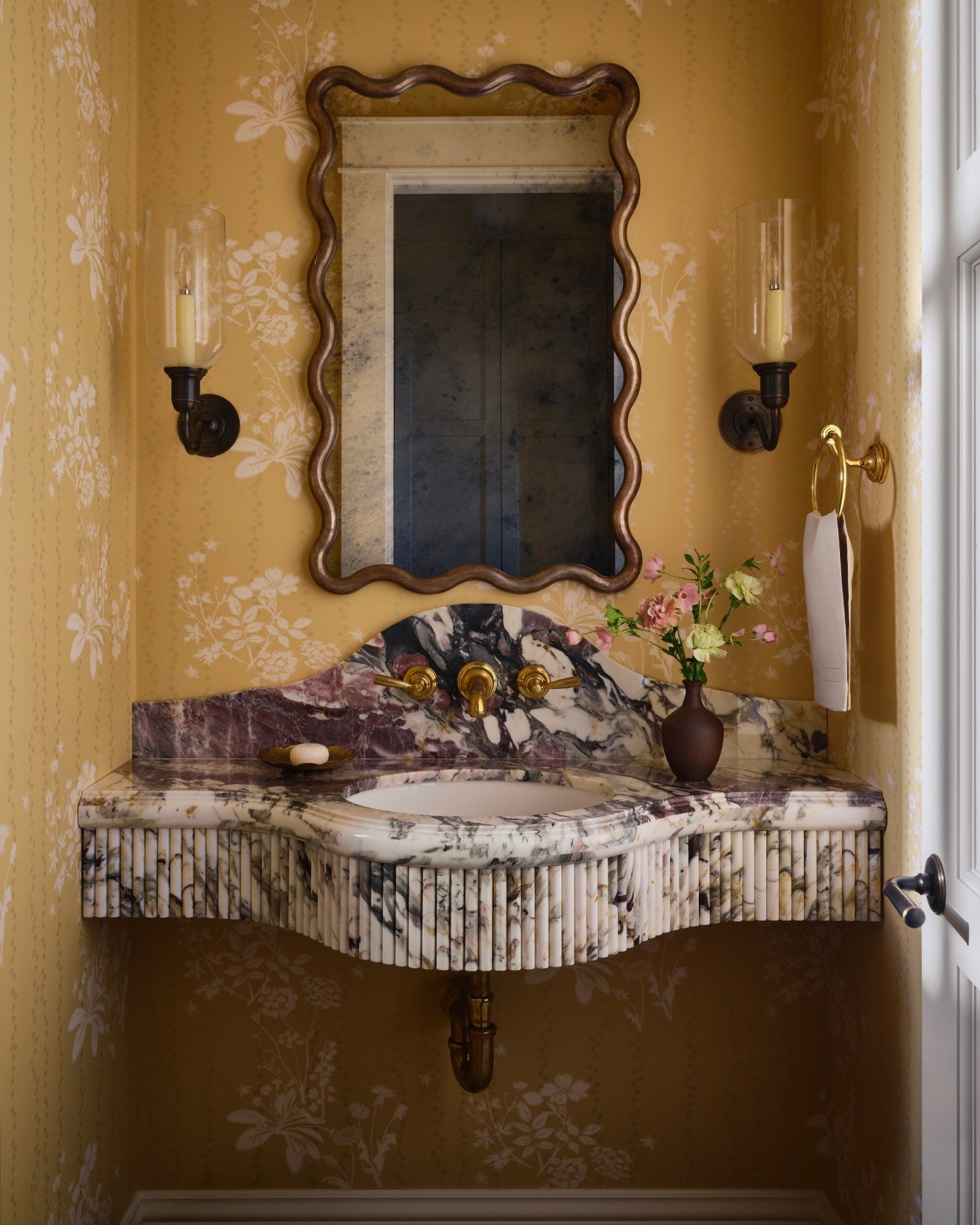 Vintage bathroom with floral yellow wallpaper, a marble sink with brass fixtures, a mirror with a wavy frame, wall sconces, a vase with flowers, and a towel holder.