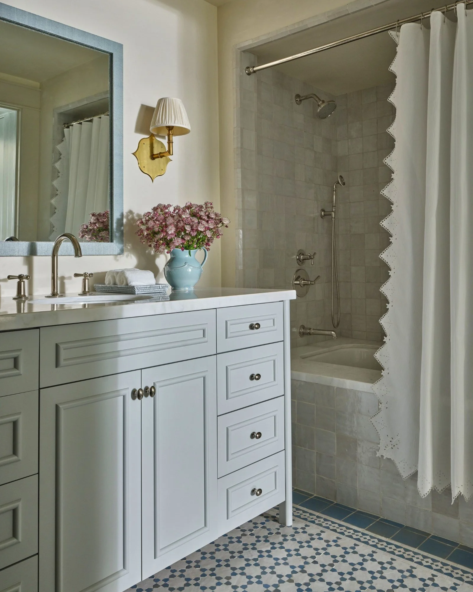 A bathroom with a white vanity, a large mirror with a light blue frame, a vase with pink flowers, a wall-mounted lamp, and a shower with a white curtain, beige tiled walls, and blue mosaic tiled floor.