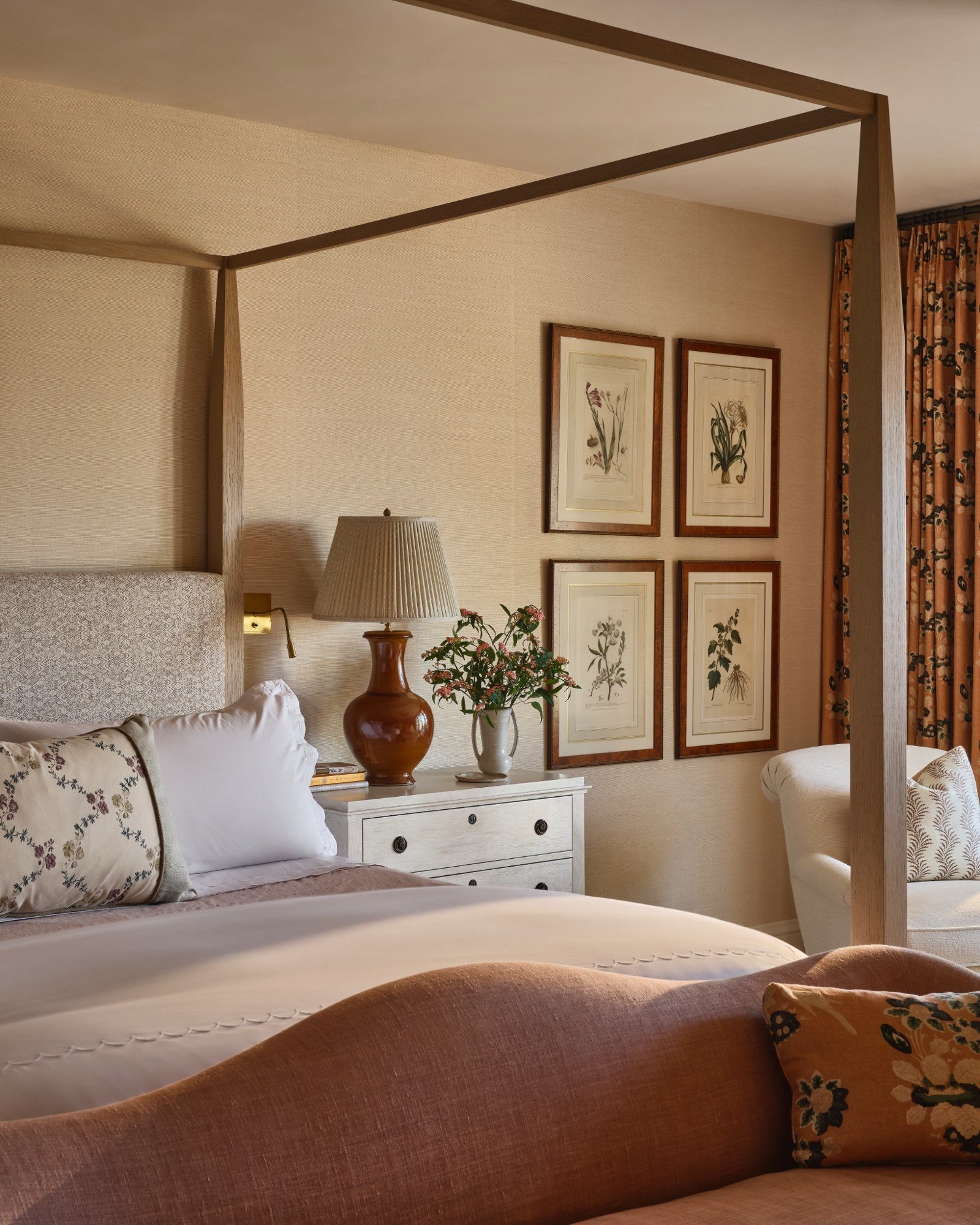 A cozy bedroom with a four-poster bed, a white bedside table with a brown lamp and a vase of flowers, framed botanical prints on the wall, and a window with floral curtains.