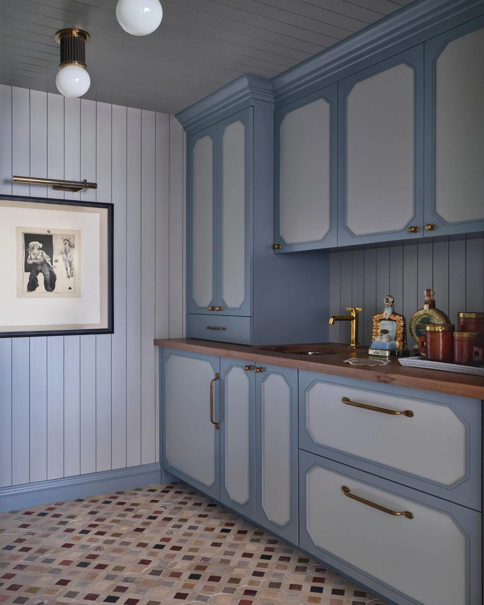 A kitchen with blue cabinets, a wooden countertop, and a backsplash with vertical white paneling. There are items on the countertop including jars and a small statue. The floor has a mosaic tile pattern, and a framed black-and-white artwork is on the wall. The ceiling has a wooden finish with two round white ceiling lights.
