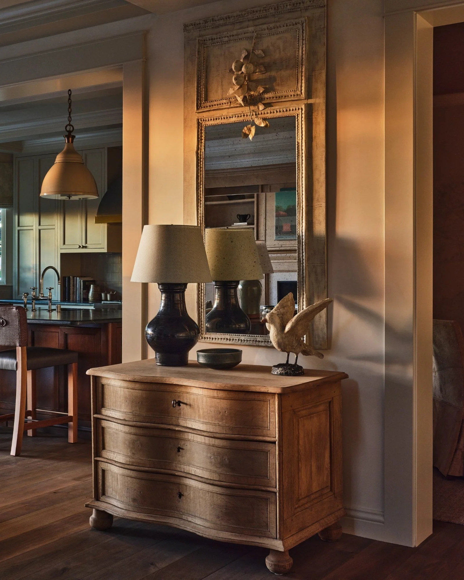 A wooden dresser with three drawers, topped with two dark lamps with beige shades, a small bowl, and a bird sculpture. Behind it hangs a large decorative mirror and a branch with leaves. Part of a kitchen and dining area is visible in the background.