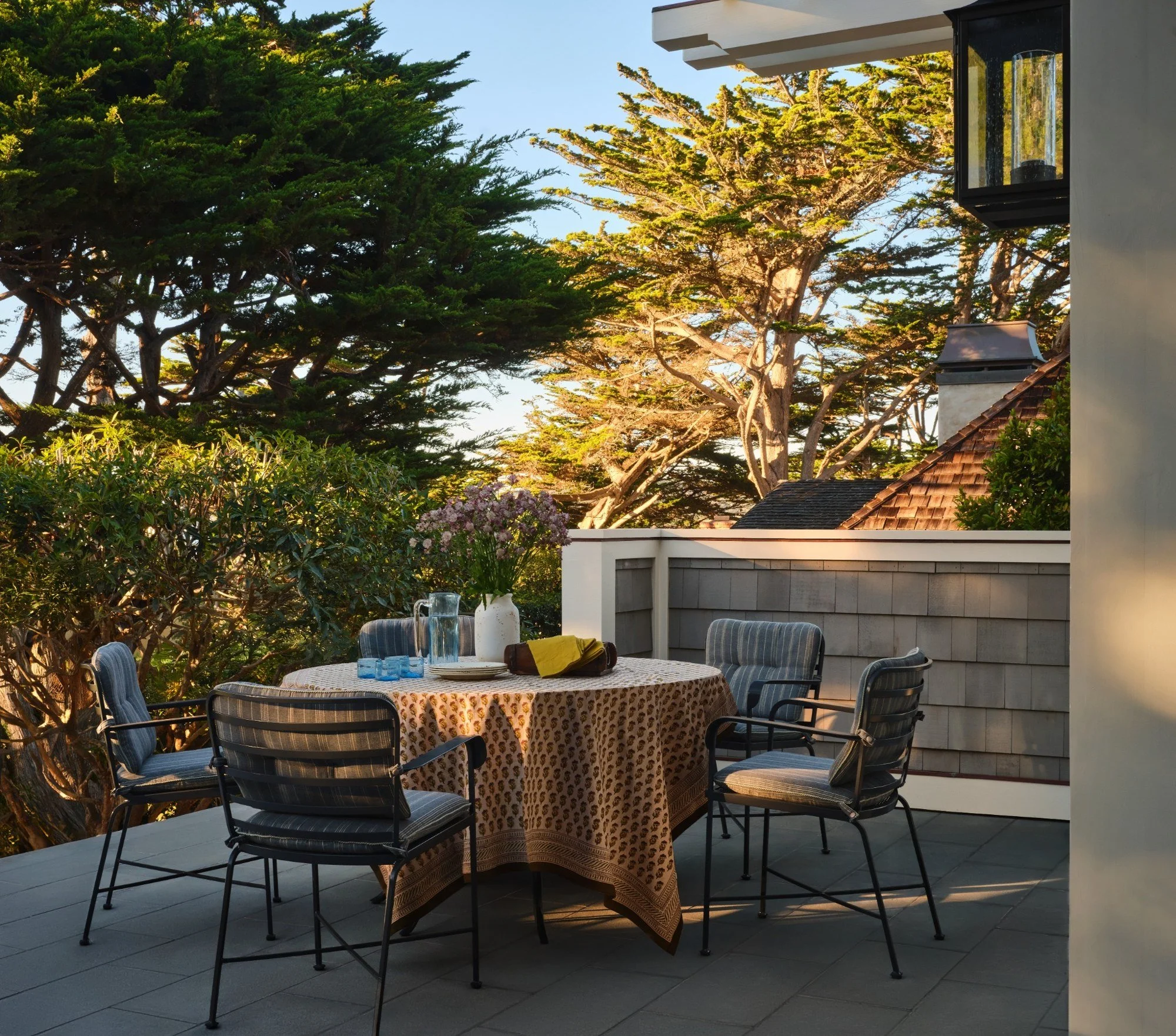 Outdoor patio with a round table covered with a patterned tablecloth, surrounded by four chairs, set with a vase of flowers, water glasses, pitchers, plates, and napkins, with trees and sky in the background.