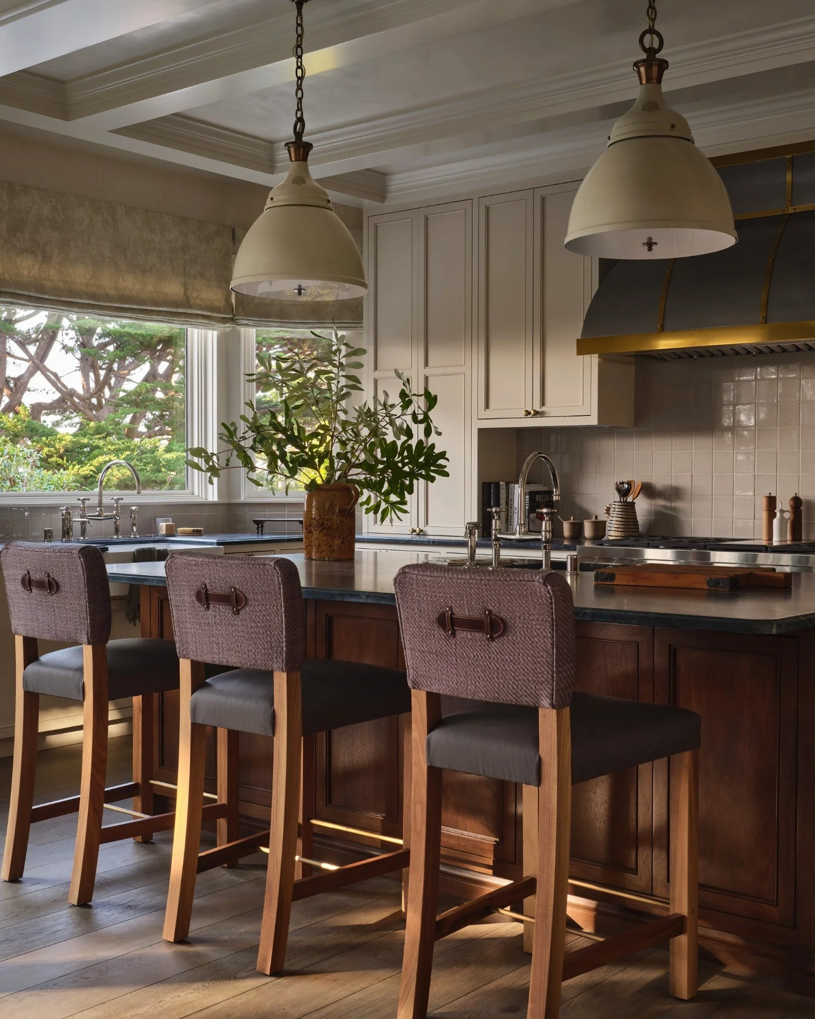 A kitchen with white upper cabinets, a dark countertop, three bar stools with upholstered seats, a large window with a view of trees, two hanging pendant lights, and a potted plant on the counter.
