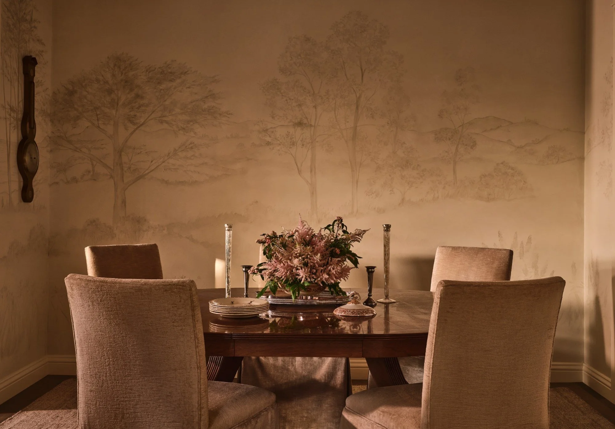 A dining room with a round wooden table surrounded by six beige upholstered chairs. The table has a floral centerpiece in shades of pink and purple, with six candlesticks and plates. The background wall features a mural of trees and a landscape in muted tones.