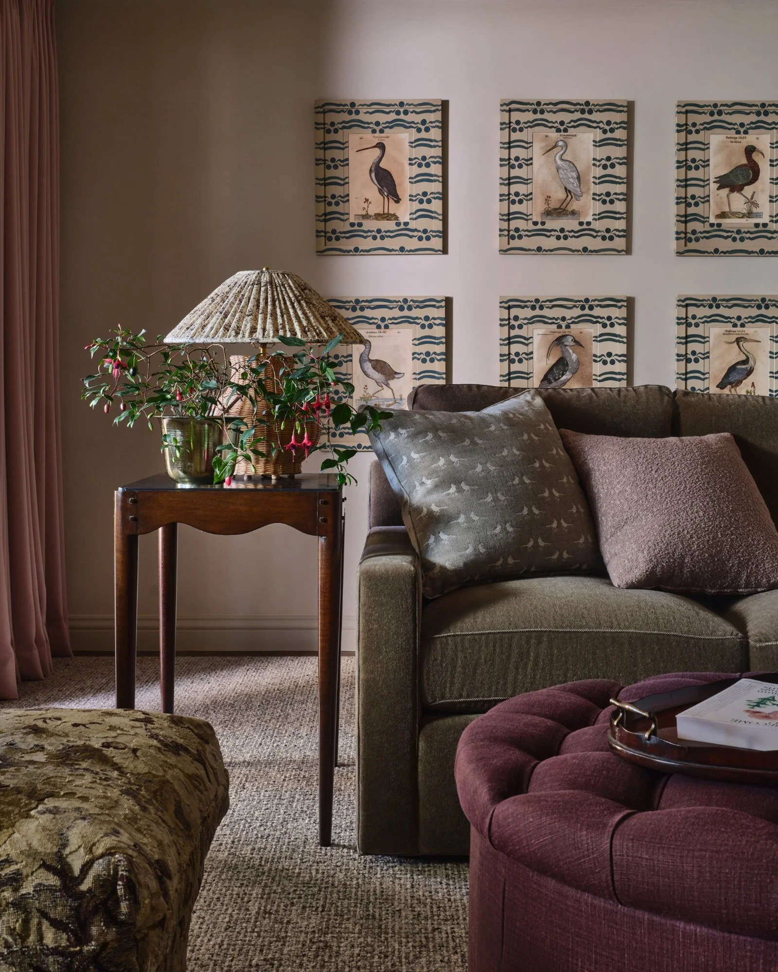 A cozy living room with framed bird illustrations on the wall, a wooden side table with a flower arrangement and a lamp, a green sofa with pillows, and an upholstered armchair with a tray holding a book.