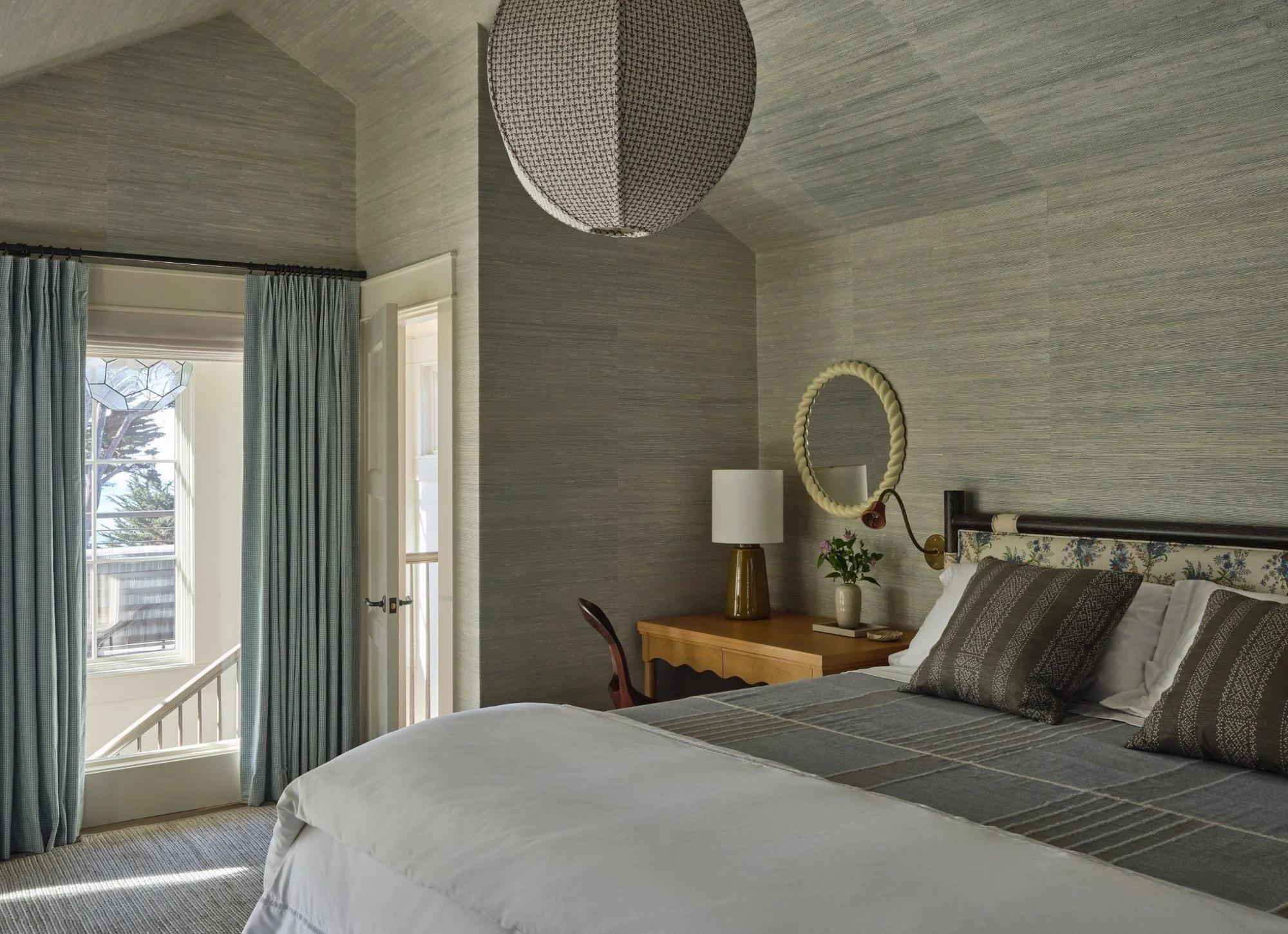 A cozy bedroom with a bed, pillows, a bedside table with a lamp, a vase with flowers, a mirror, and a window with curtains revealing a view outside.