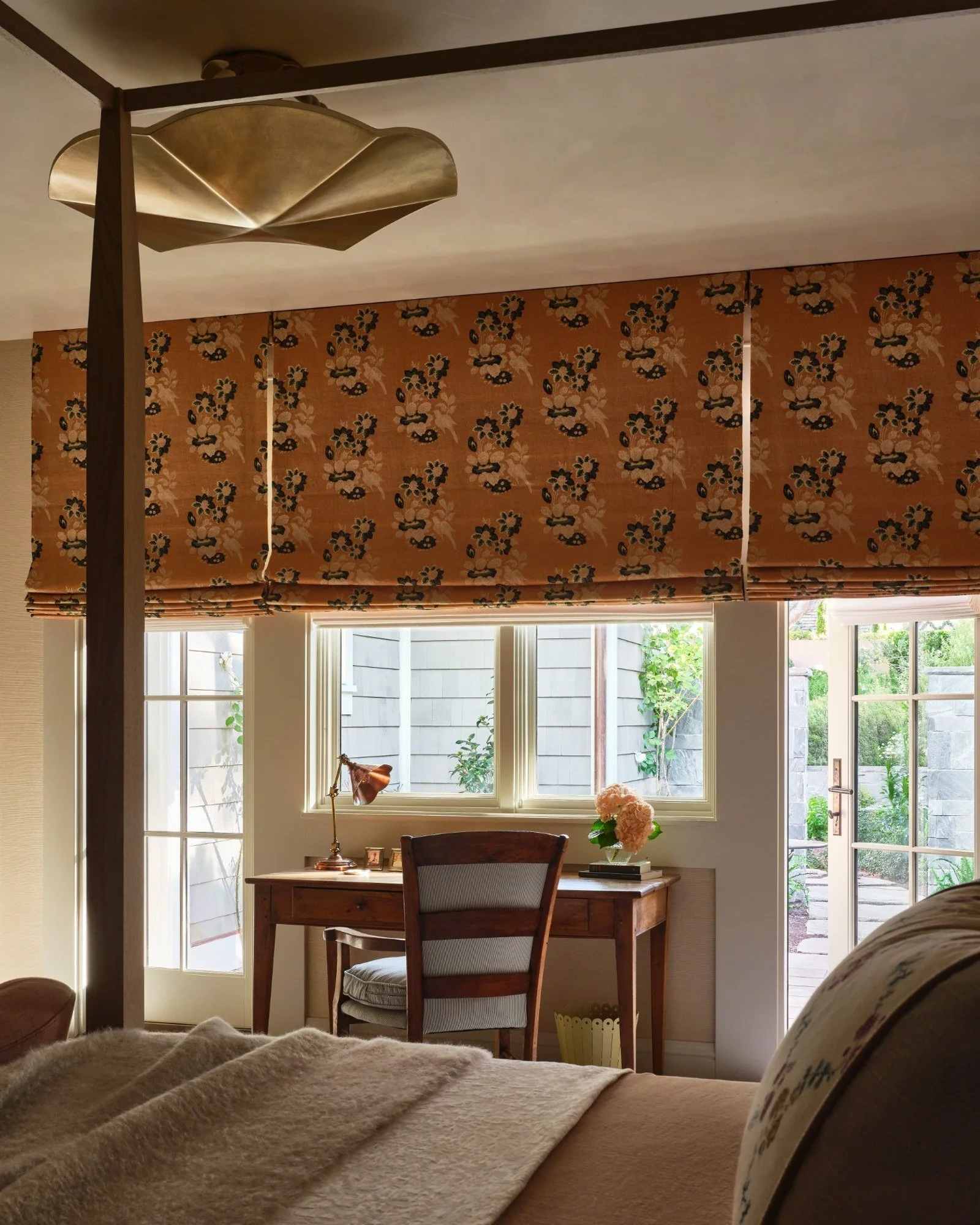 View of a cozy bedroom with a desk by a window, a wooden chair, a pink flower bouquet, a desk lamp, and an outdoor garden visible through the window and door, with a floral-patterned valance and a bed in the foreground.