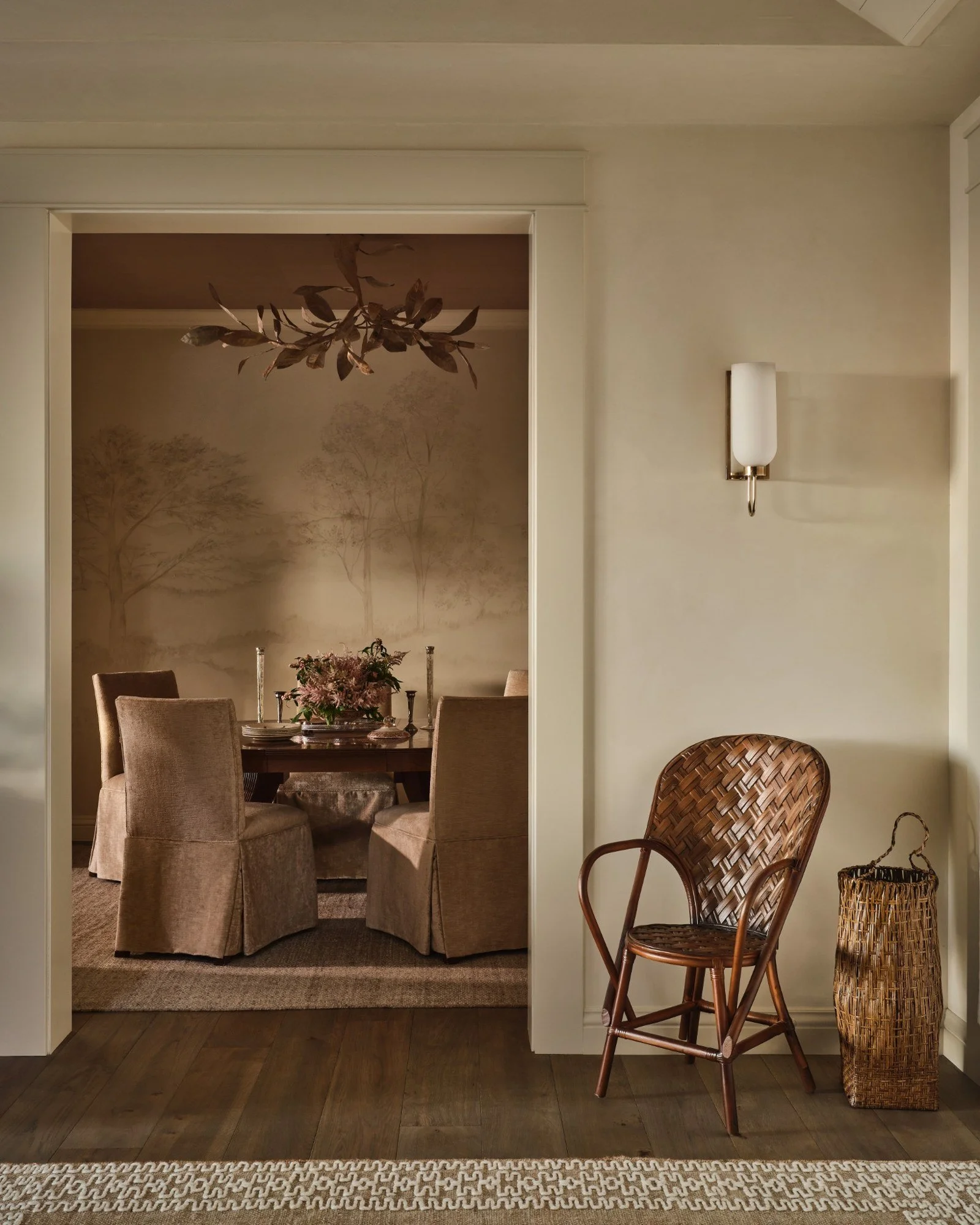 Living room with wicker basket and wooden chair, doorway leading to a dining area with a round table and six upholstered chairs, floral centerpiece, wall sconces, and tree mural in the background.