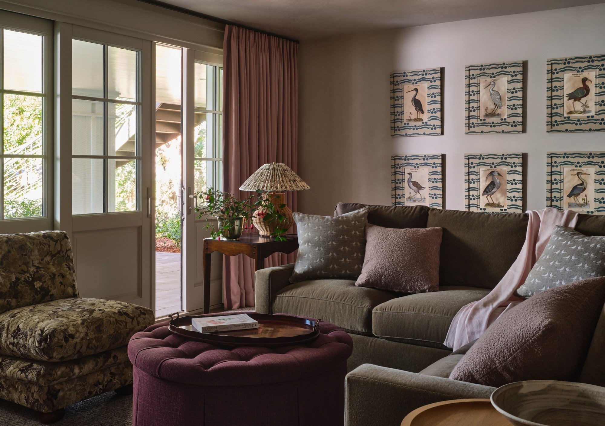 Living room with large windows, pink curtains, a beige sofa with patterned pillows, a floral armchair, a round burgundy ottoman with a tray and books, a side table with a lamp and a potted plant, and framed bird illustrations on the wall.