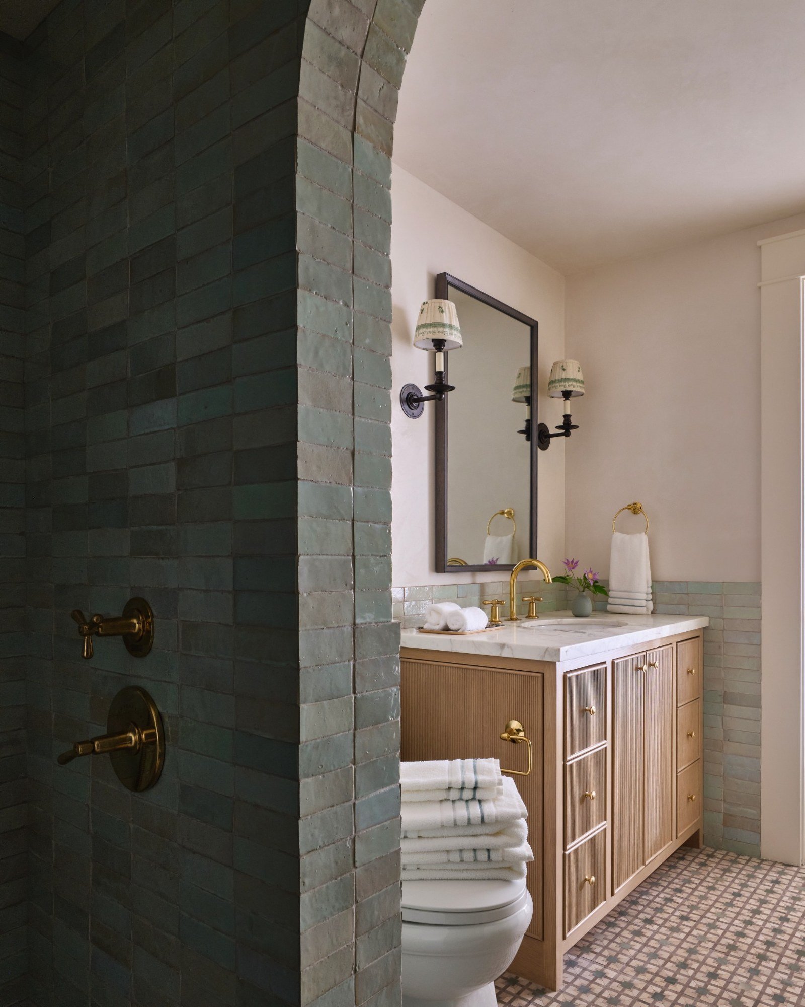 Bathroom with a wooden vanity, marble countertop, gold fixtures, mirror, wall sconces, towel rack, folded towels, toilet, and tiled shower area with brass fixtures.