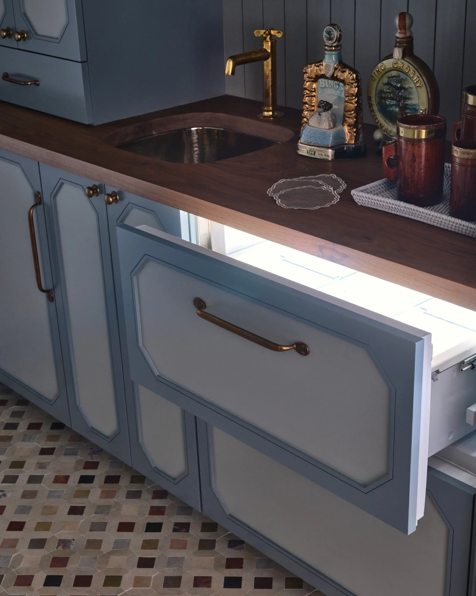A kitchen countertop with a blue cabinet drawer partially open, showing a wooden countertop, small decorative dishes, bottles, a brass faucet, and a dark-colored backsplash.