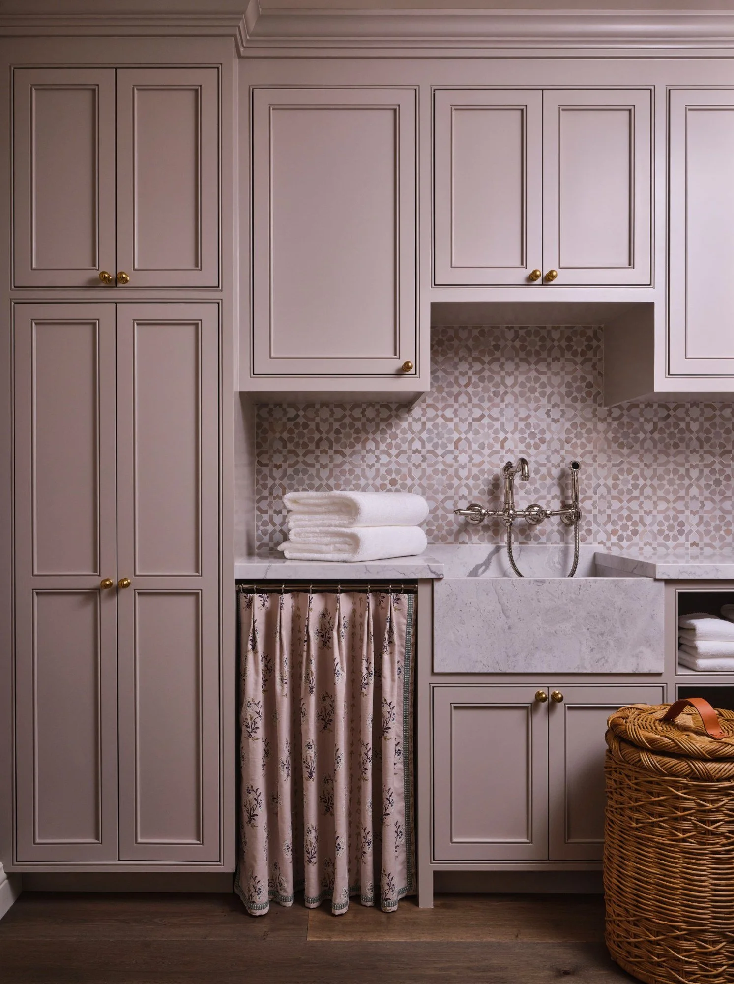 View of a laundry or utility room with beige cabinets, white towels, a decorative curtain covering a small cabinet, and a wicker laundry basket.