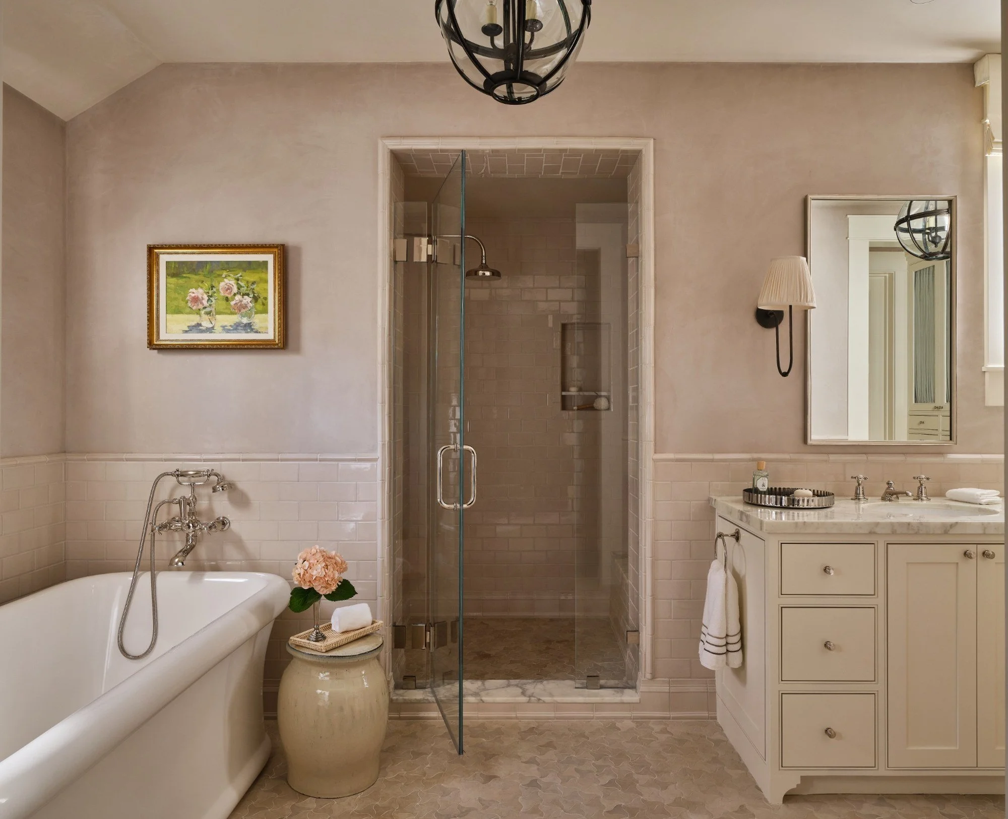 A bathroom with a white bathtub, a small table with pink flowers, a glass-enclosed shower, a cream-colored vanity with a mirror, a wall-mounted lamp, and a framed floral painting.
