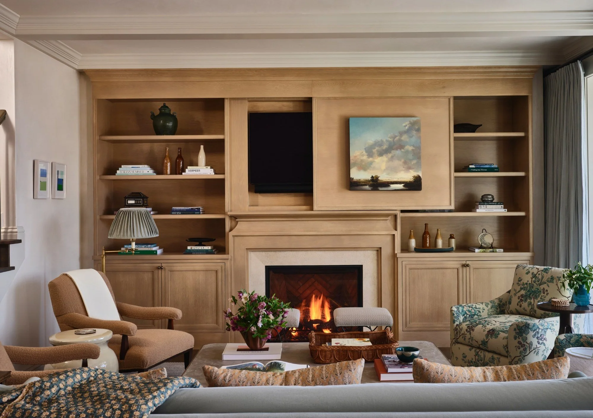 Living room with wood-paneled wall, built-in shelves, a fireplace with a fire, a framed landscape painting, a beige armchair, a floral upholstered sofa, a table with a flower arrangement, books, and decorative items.