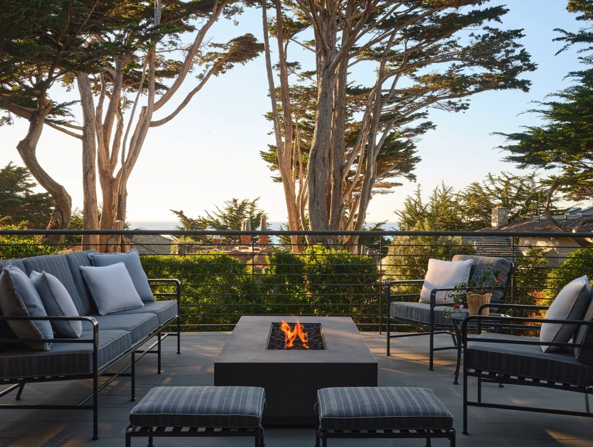 Outdoor patio with two striped sofas and two matching ottomans around a fire pit, surrounded by trees and a house with a sloped roof in the background.