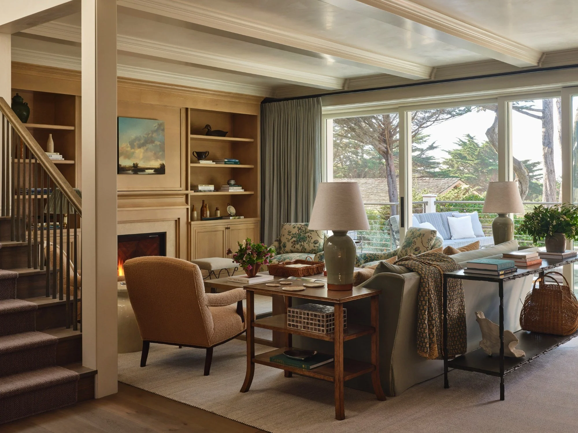 Living room with beige and floral furniture, large windows showing trees outside, fireplace, built-in wooden shelving, lamps, and decorative objects.