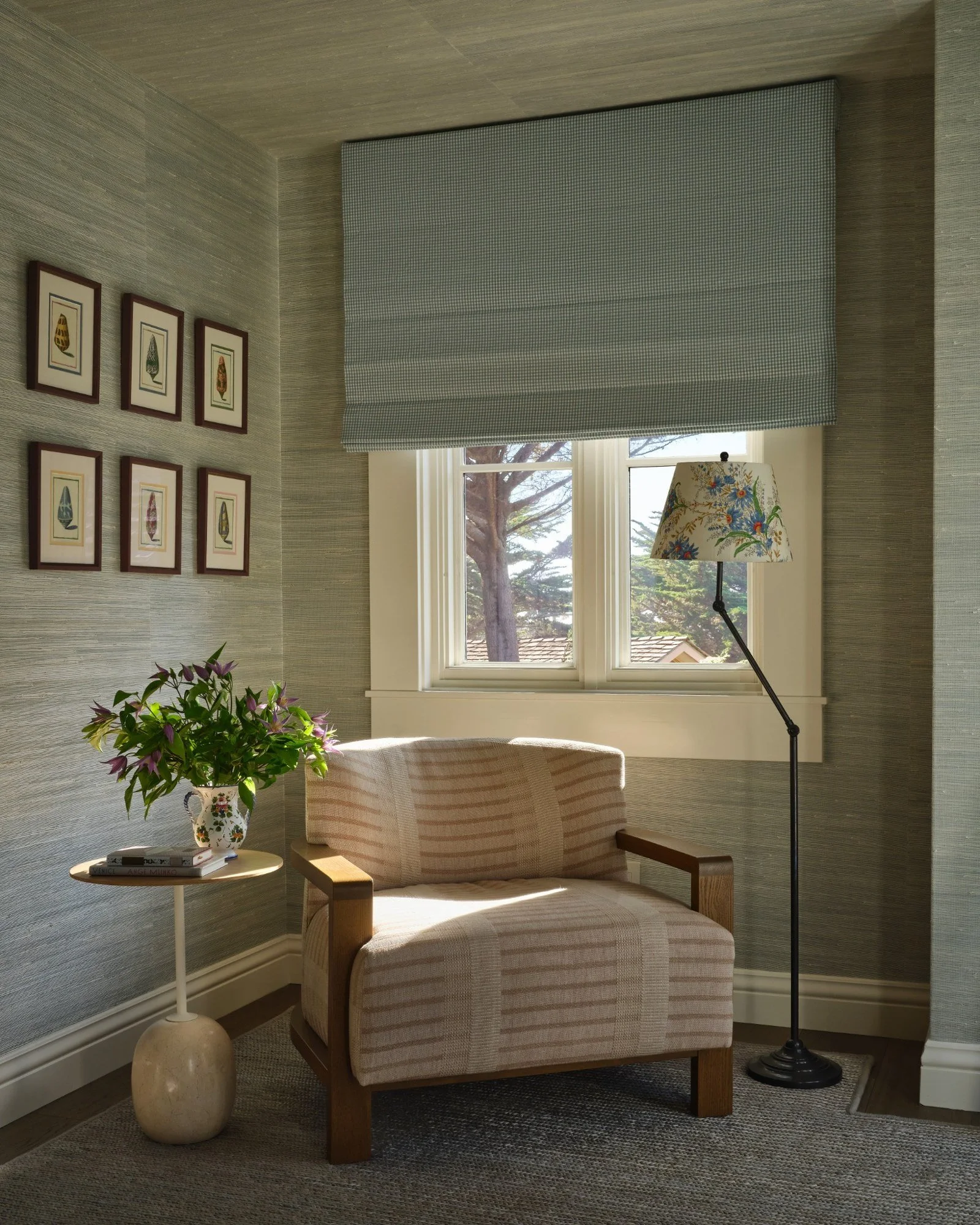 Cozy corner with a pink striped armchair, a side table with flowers, a standing lamp with a floral lampshade, and wall art of framed nature illustrations.