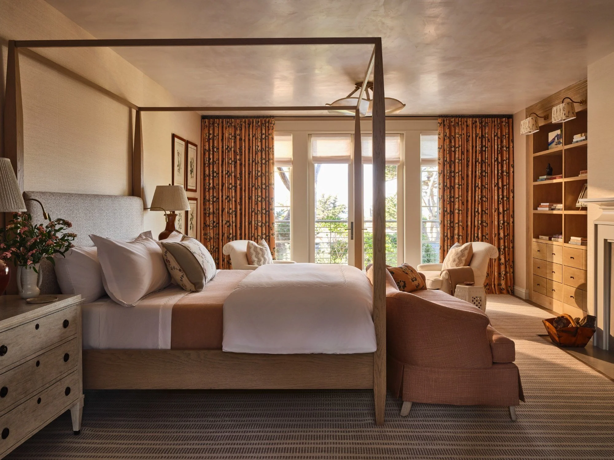 A bedroom with a four-poster bed, beige bedding, a pink armchair, and large windows with orange floral curtains.