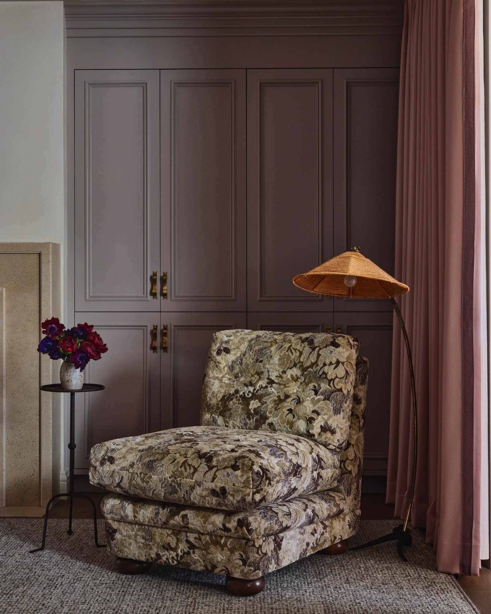 A cozy corner with a floral upholstered armchair, a small side table with a vase of flowers, a floor lamp with a woven shade, a large wardrobe with paneled doors, pink curtains, and a textured carpet.