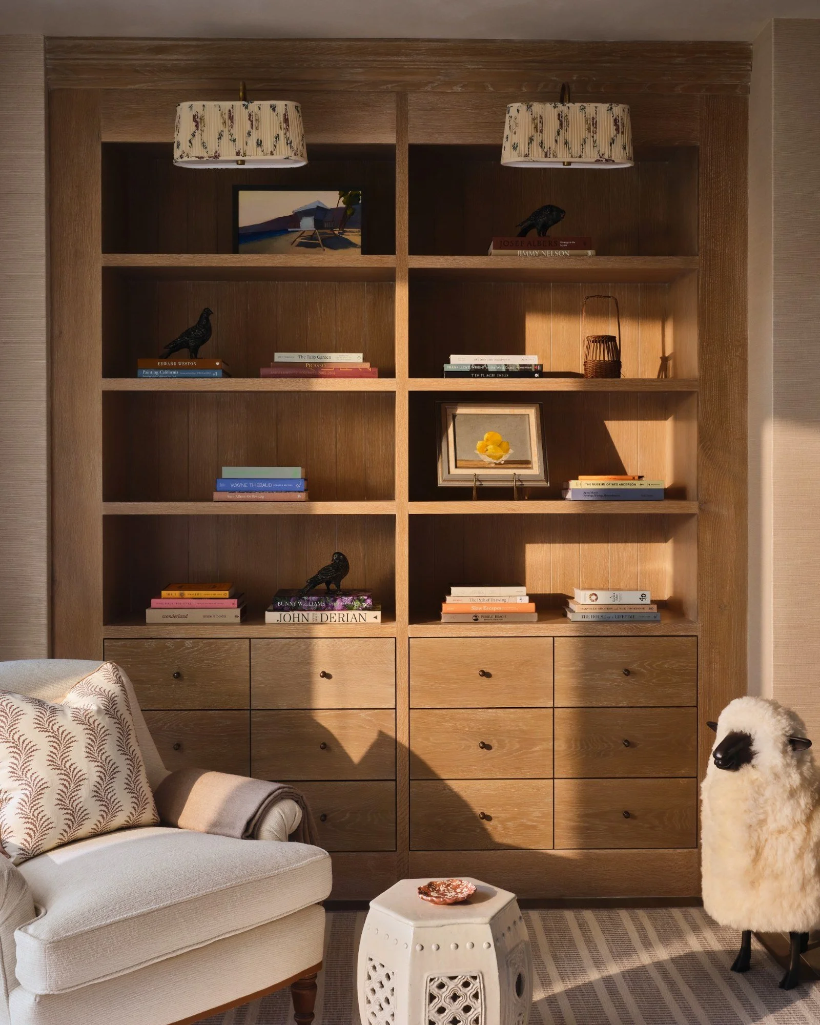 A wooden bookshelf in a living room with decorative items, books, and a framed picture. There is a beige armchair with a patterned pillow, a small white ceramic table, and a stuffed sheep toy to the right.