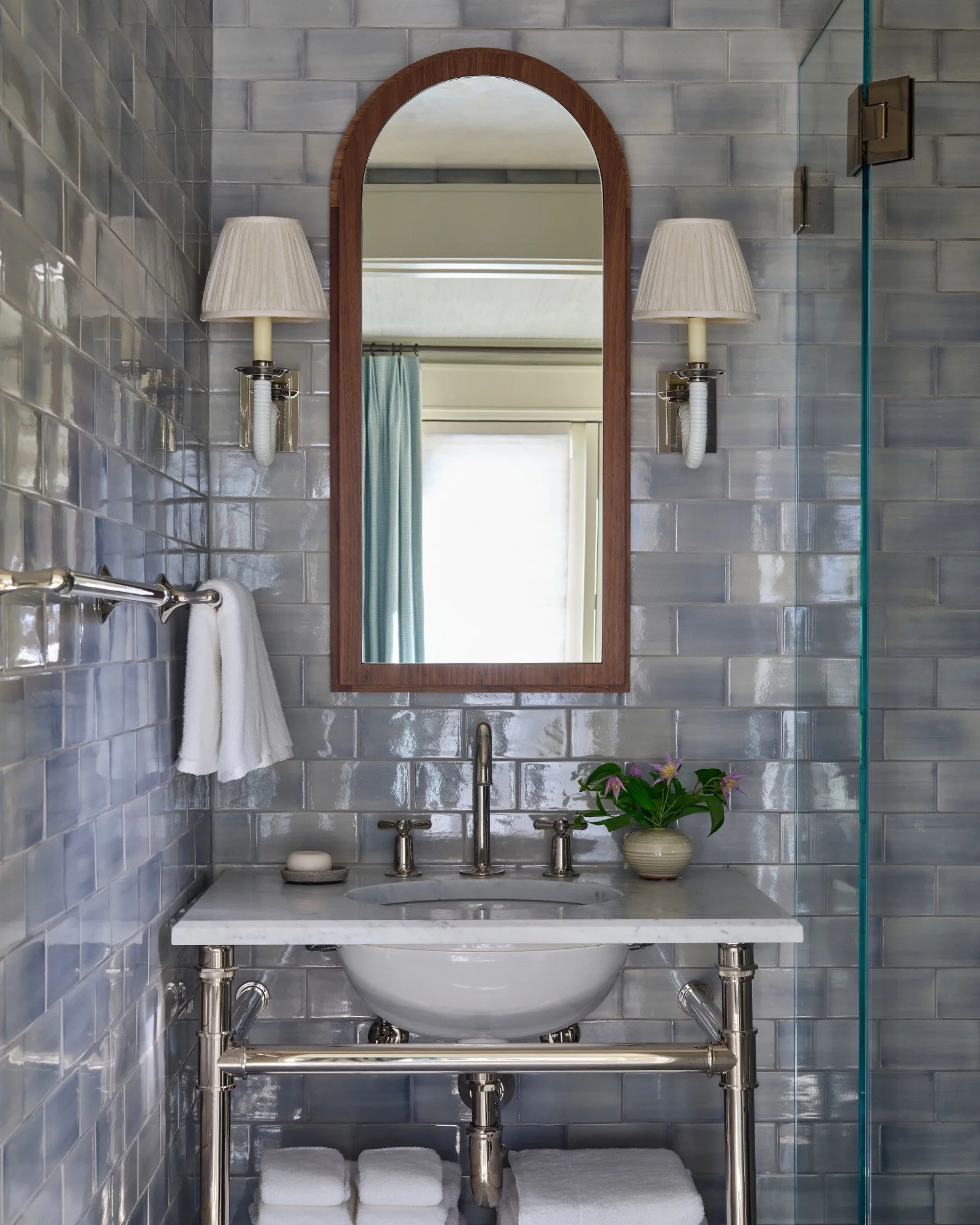 Bathroom with gray tiled walls, a marble countertop sink, a mirror with a wooden frame, two wall-mounted lamps with pleated shades, a towel rack with white towels, a potted plant, and soap on the counter.