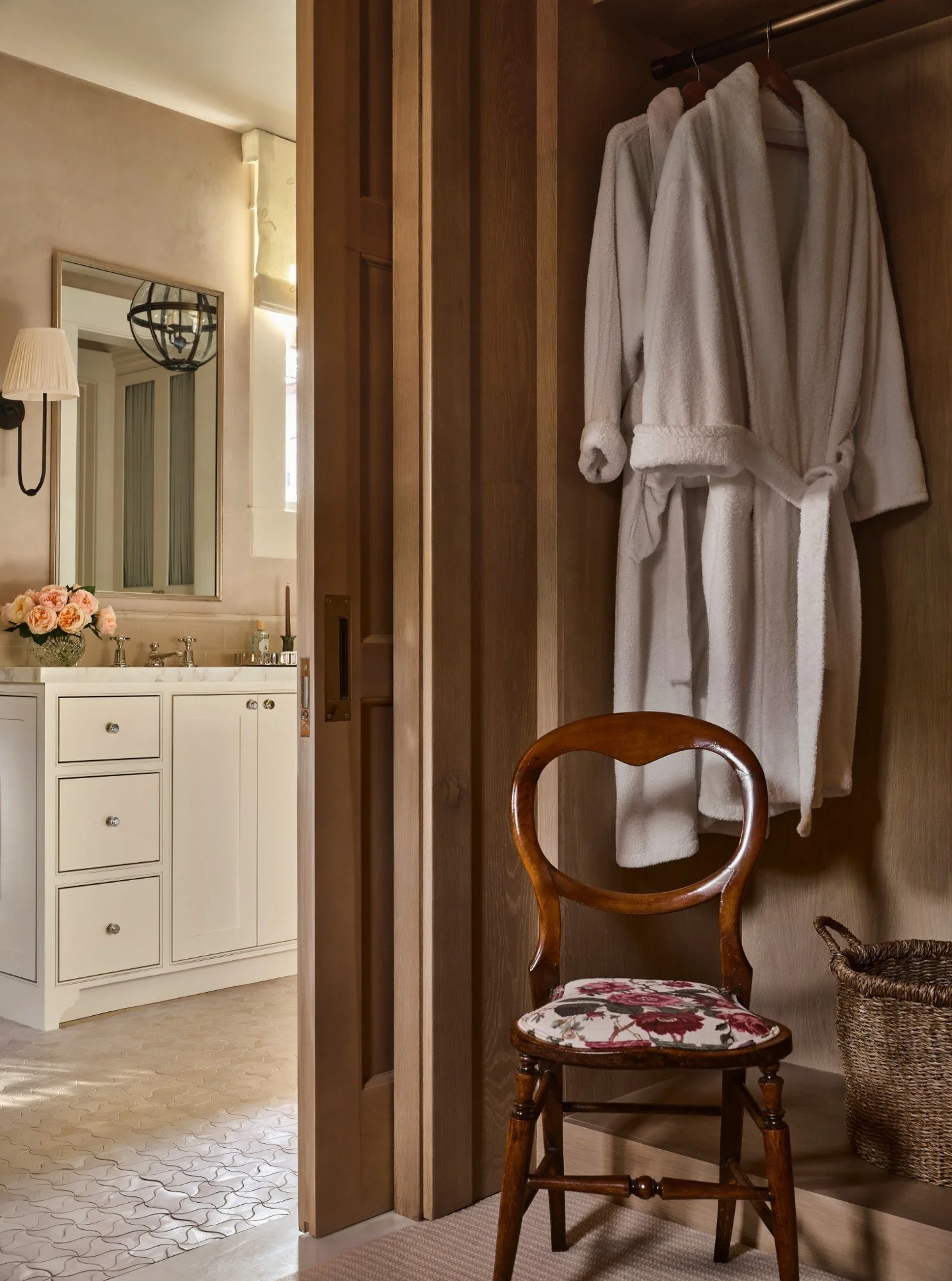 A wooden closet with a white bathrobe hanging inside, a vintage chair with a floral cushion, a laundry basket, a bathroom with a vanity, mirror, and flowers in the background.