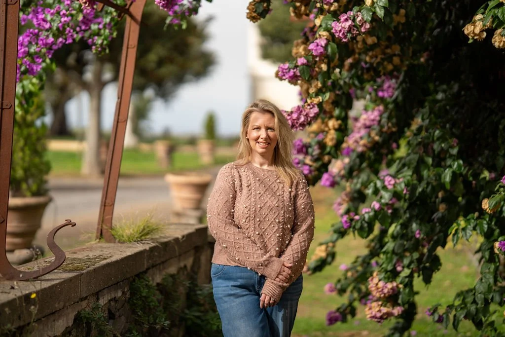 A woman with blonde hair smiling in front of blooming pink and purple flowers outdoors at an Italian countryside villa.