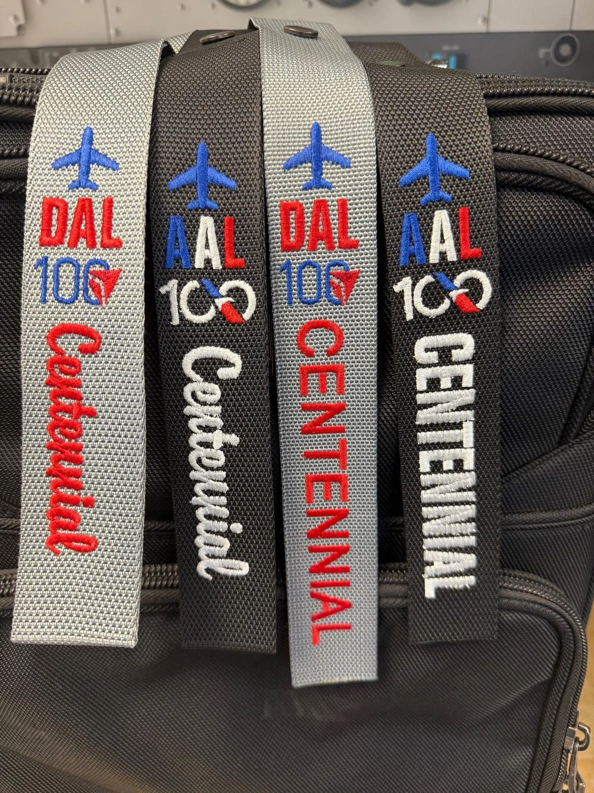 Four embroidered luggage tags in black and white colors with blue airplane symbols and red, white, black, and blue text, attached to a gray textured bag.