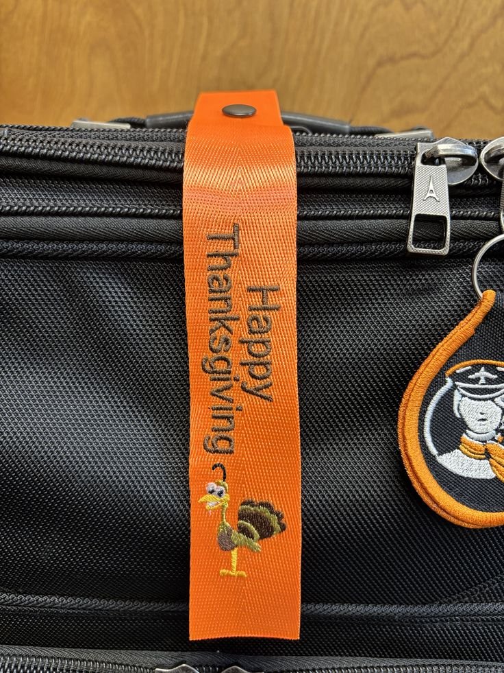 Close-up of a black travel bag with an orange luggage tag that has the text 'Traveling With Thanksgiving' and an embroidered turkey image. Part of a circular embroidered patch featuring a smiling character wearing a pilot's hat is visible on the right side of the bag.