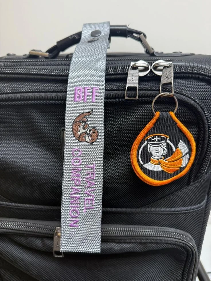 Close-up of a black travel bag with two zippers, a luggage tag with a gray strap embroidered with 'BFF' and a cat, and a circular keychain with a pig in a graduation cap and gown.