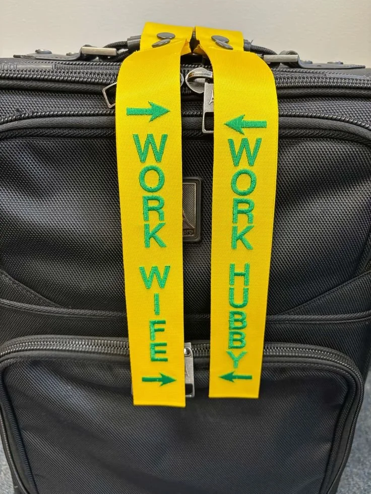 A black rolling suitcase with yellow luggage tags hanging from the zipper. The tags have green text and directional arrows, with one reading 'WORK WIFE' and the other 'WORK HUBBY'.