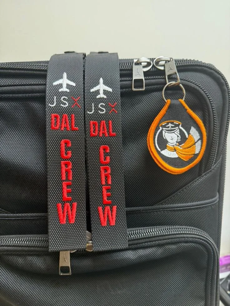 Close-up of a black suitcase with two zipper pulls, a Thimbleberry pin featuring a cartoon character in a hat and scarf, and two black luggage tags embroidered with white airplane icons, red 'DAL' text, and red 'CREW' text.