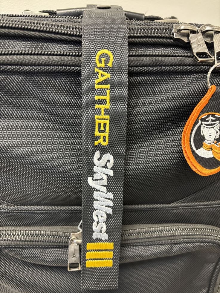 Close-up of a black luggage handle with embroidered yellow and white text that reads 'GATHER SHWAG' and a keychain featuring a stylized character wearing a mask.