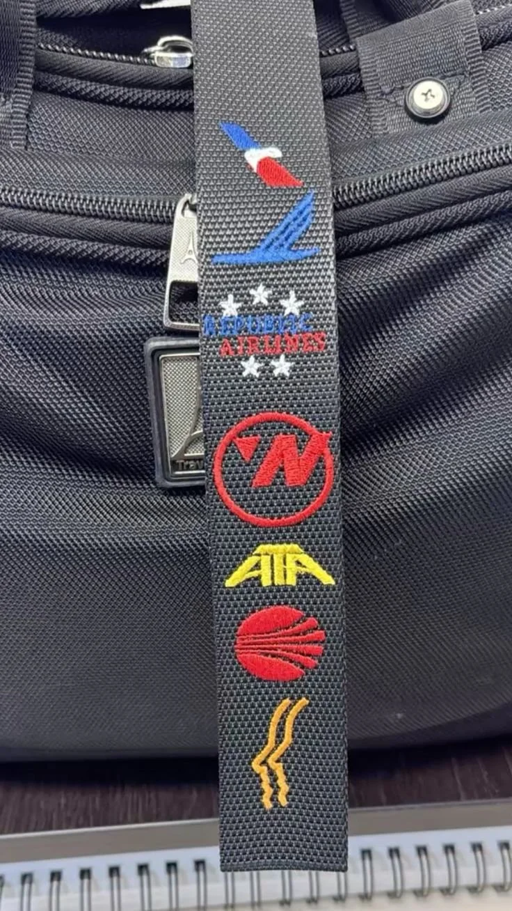 Close-up of a black fabric strap with embroidery of aviation-related logos and symbols, hanging in front of a black bag with zippers.